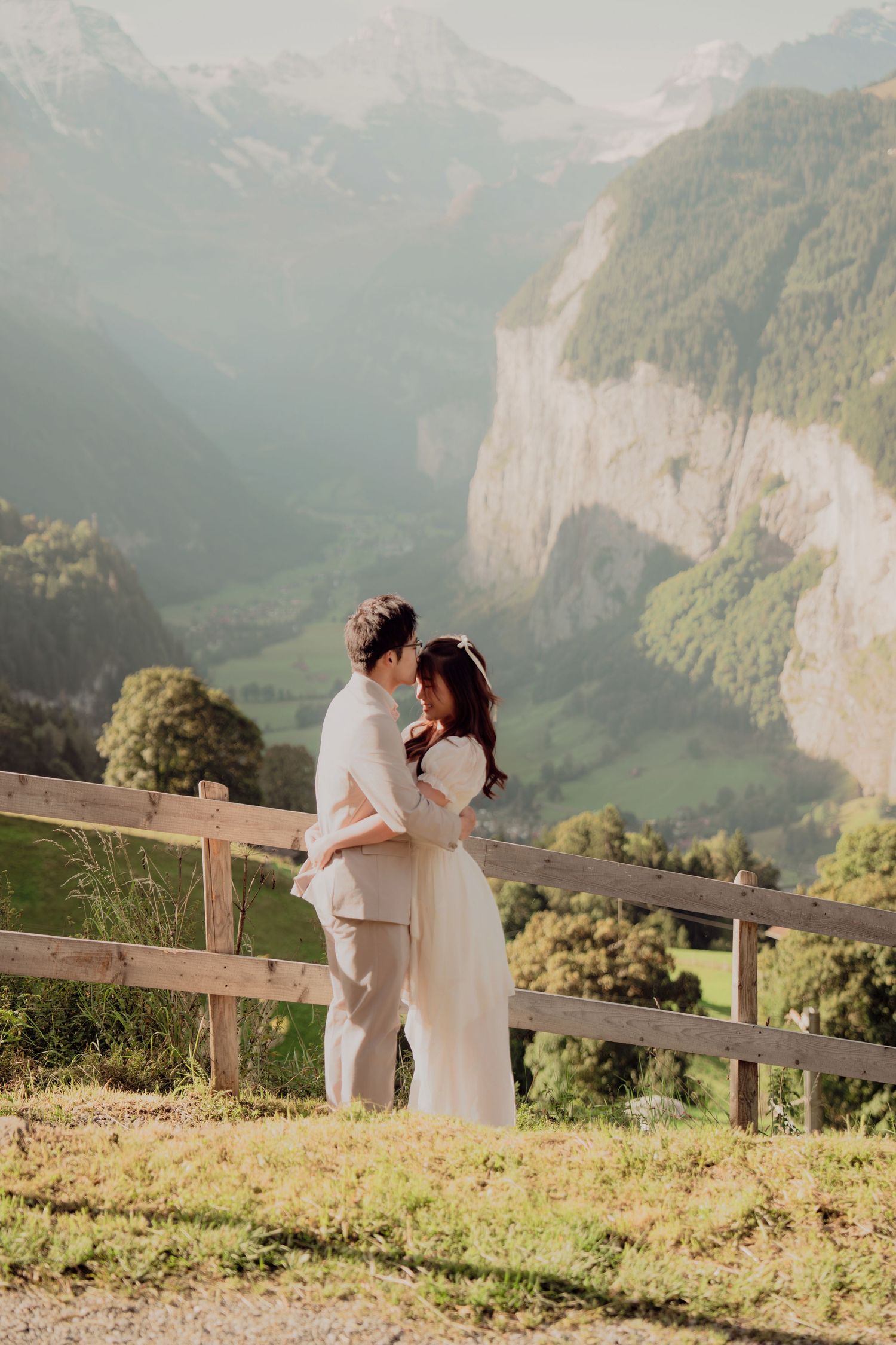 Bride and groom sharing a tender embrace by a fence overlooking a misty Swiss alpine valley.