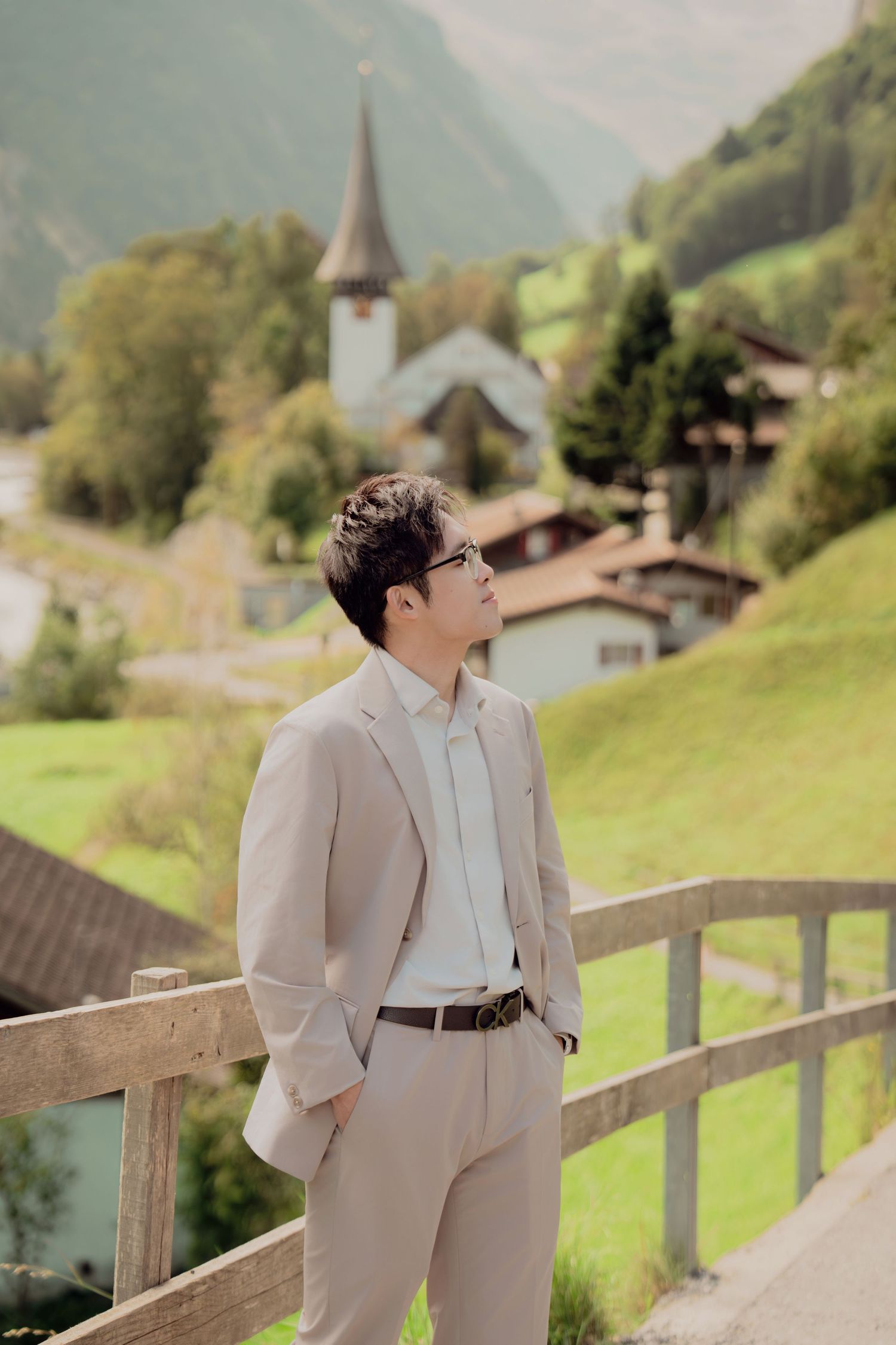 Man in beige suit standing on wooden bridge with Swiss alpine village and church steeple in background.