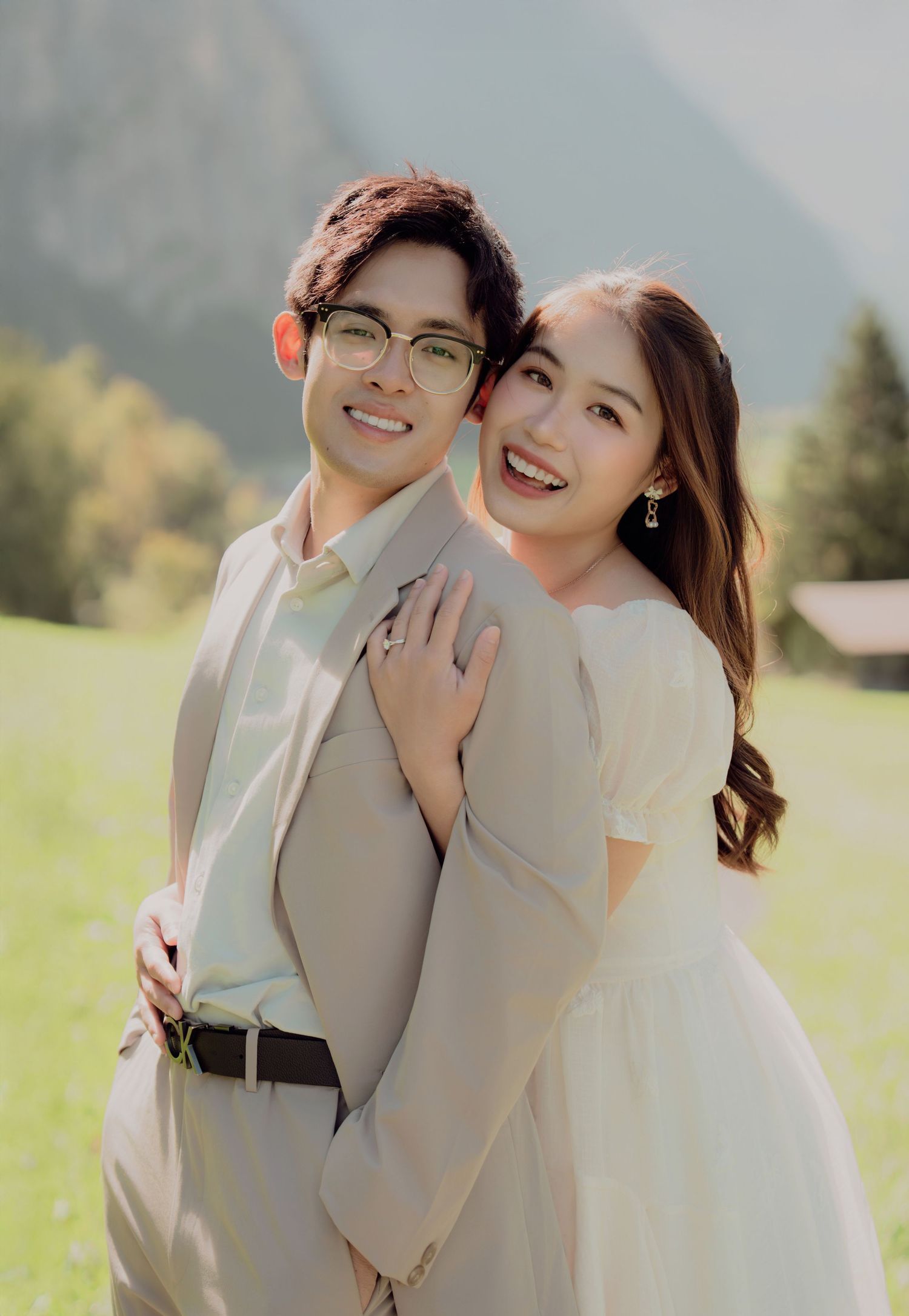 Smiling couple in beige wedding attire posing closely together with lush green Alpine mountains in the background.