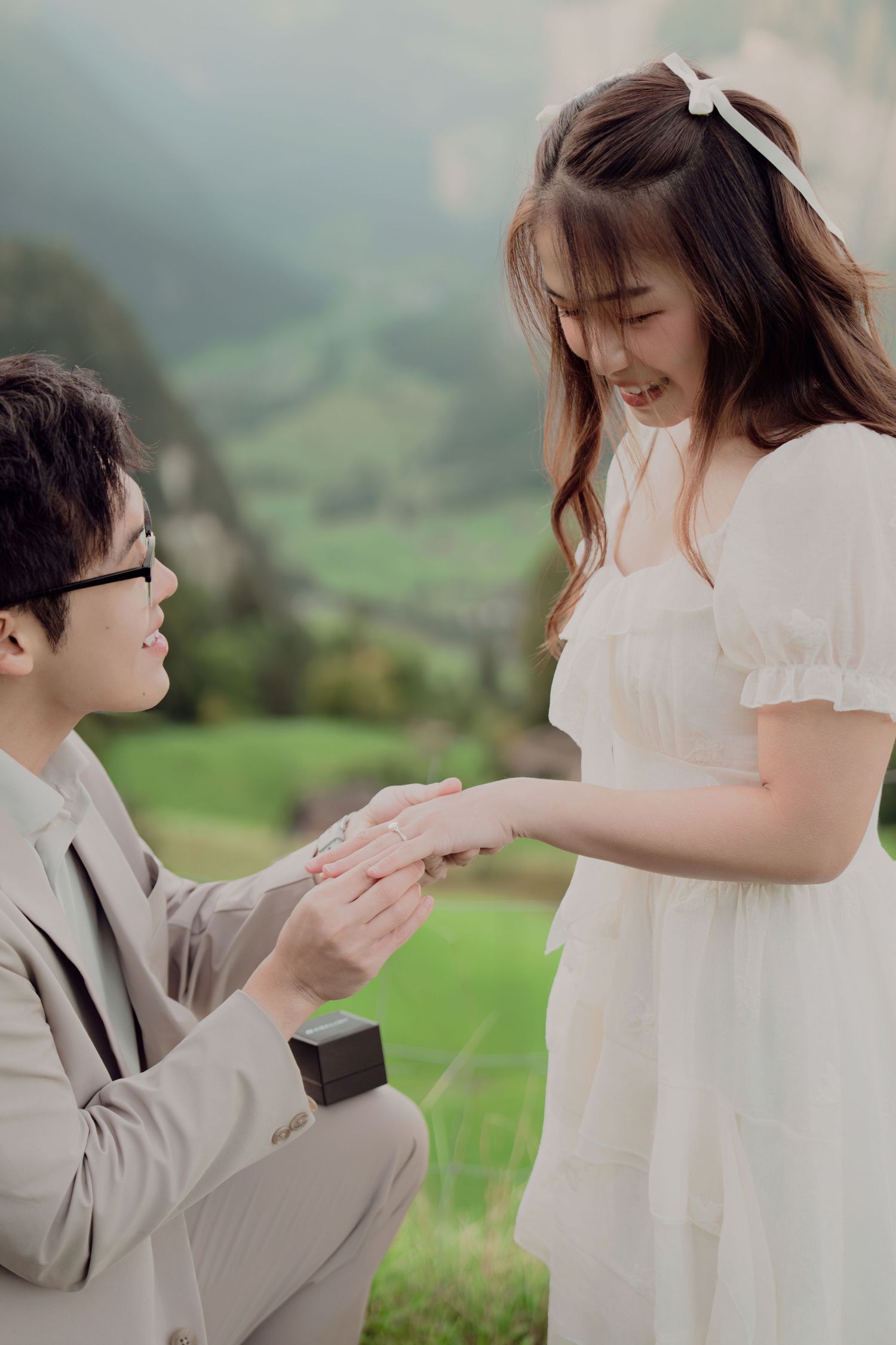 Joyful marriage proposal scene with man placing ring on woman's finger in a lush green alpine meadow.