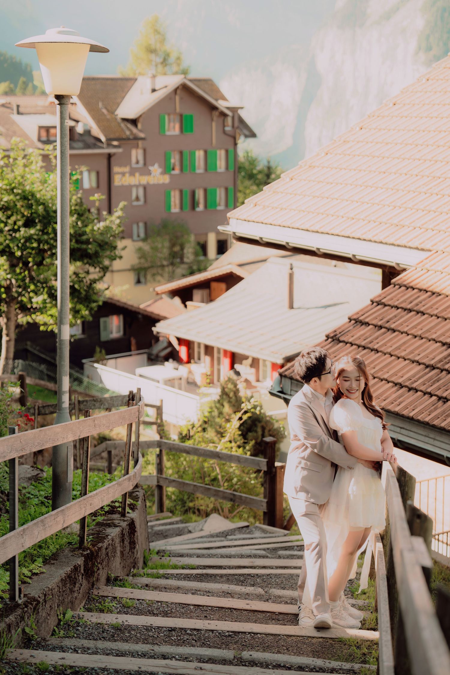 Couple embracing romantically on Swiss village staircase with traditional alpine buildings and green shutters behind.