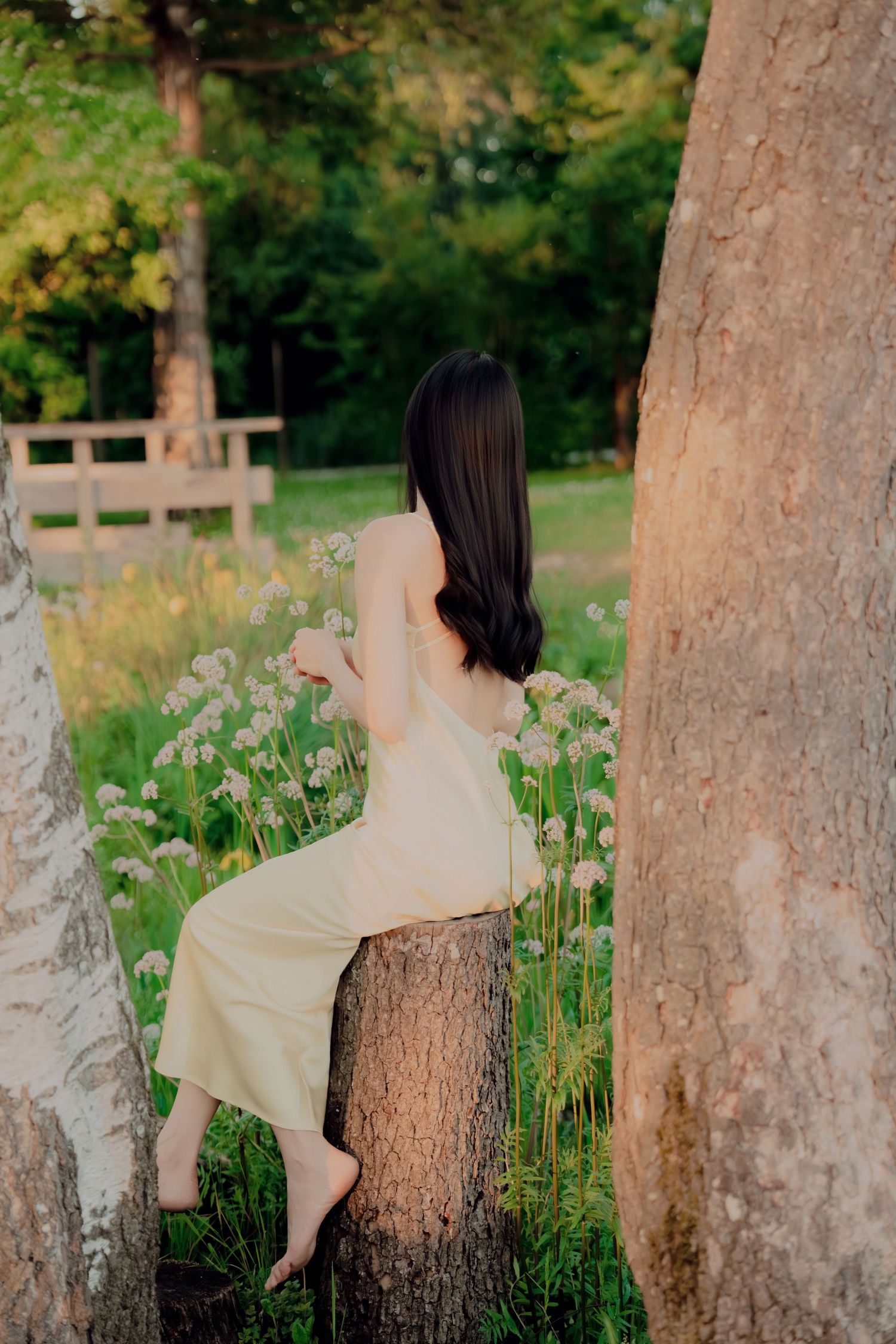Woman in yellow dress sitting barefoot on tree stump among white wildflowers in park.
