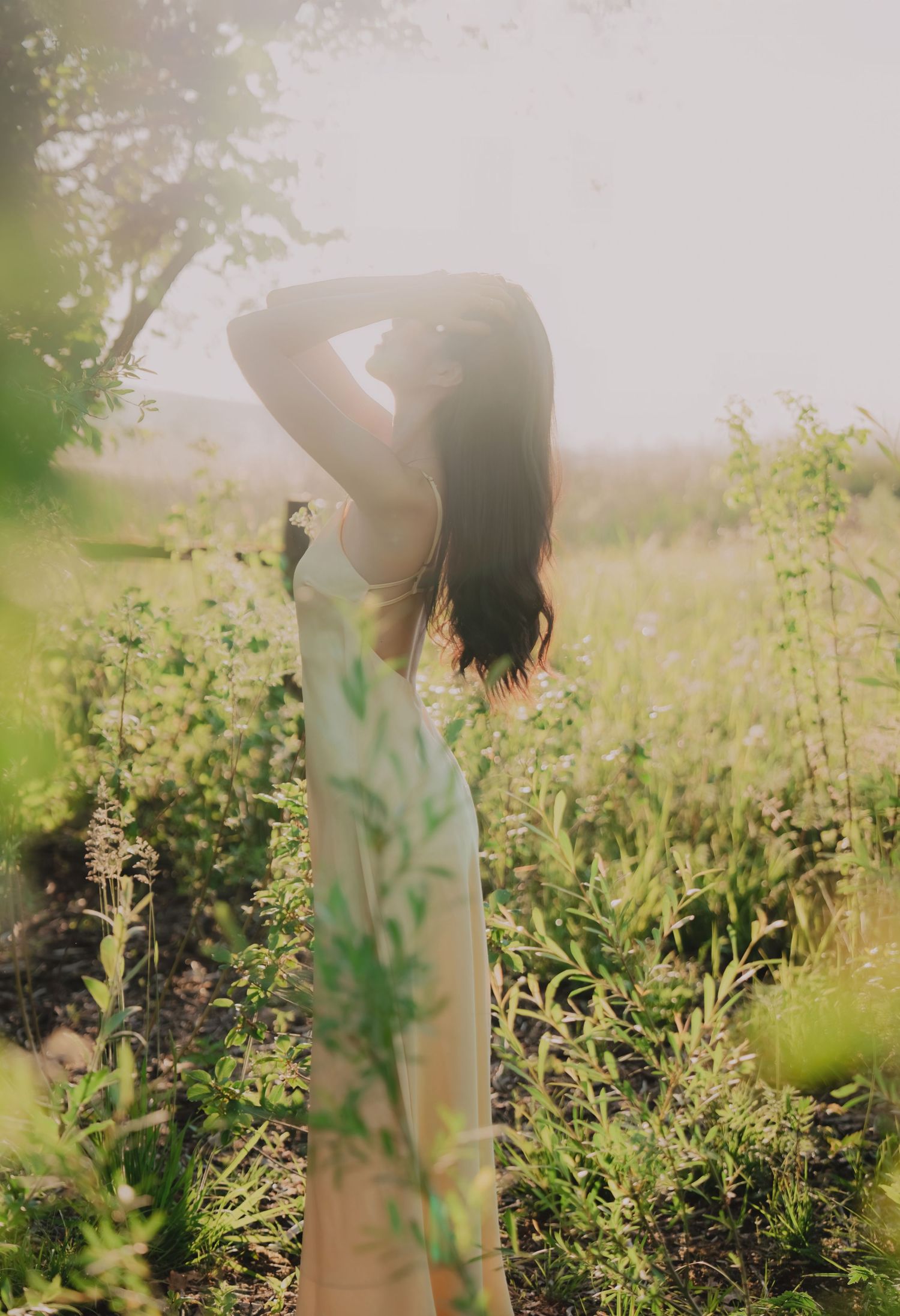 Woman in white dress standing in sunlit wildflower field with arms raised, backlit by golden sunlight.