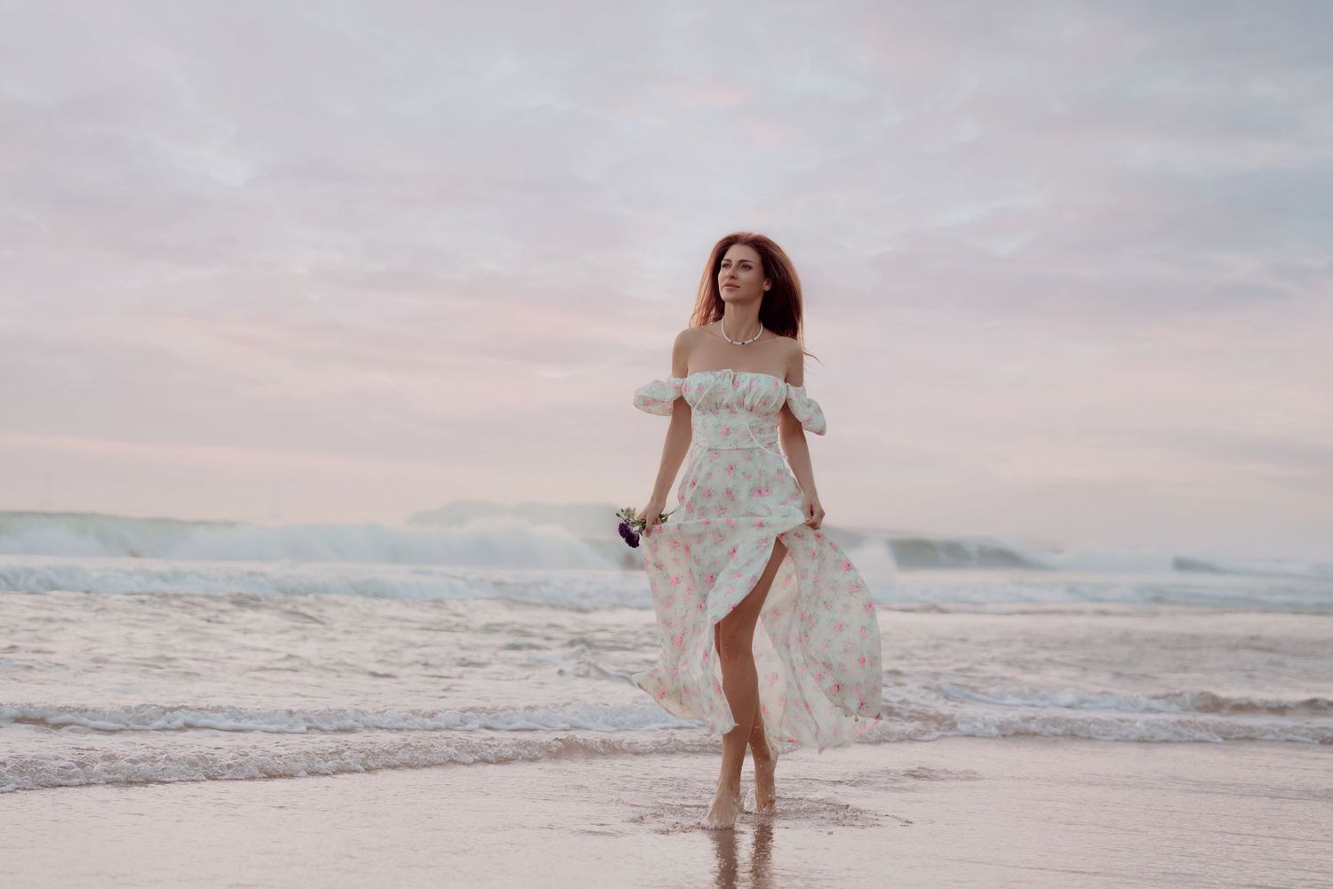 Woman in white floral off-shoulder dress walking on a beach at sunset with ocean waves in the background.