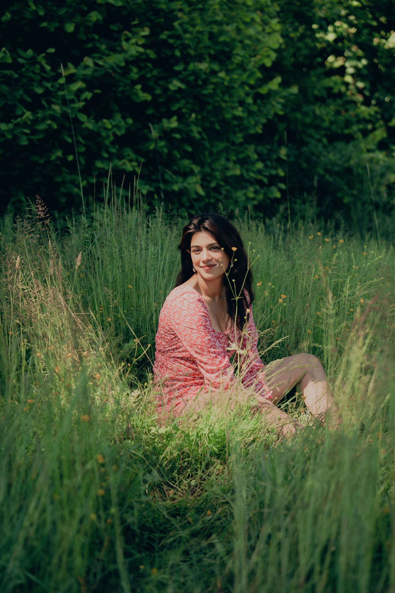 Young woman in pink dress sitting in tall green grass in a lush forest meadow, smiling at camera.