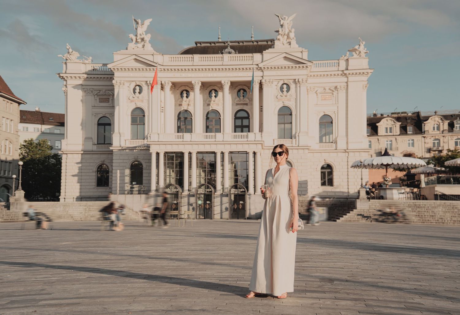 Woman in white dress standing in plaza before ornate white neoclassical opera house.