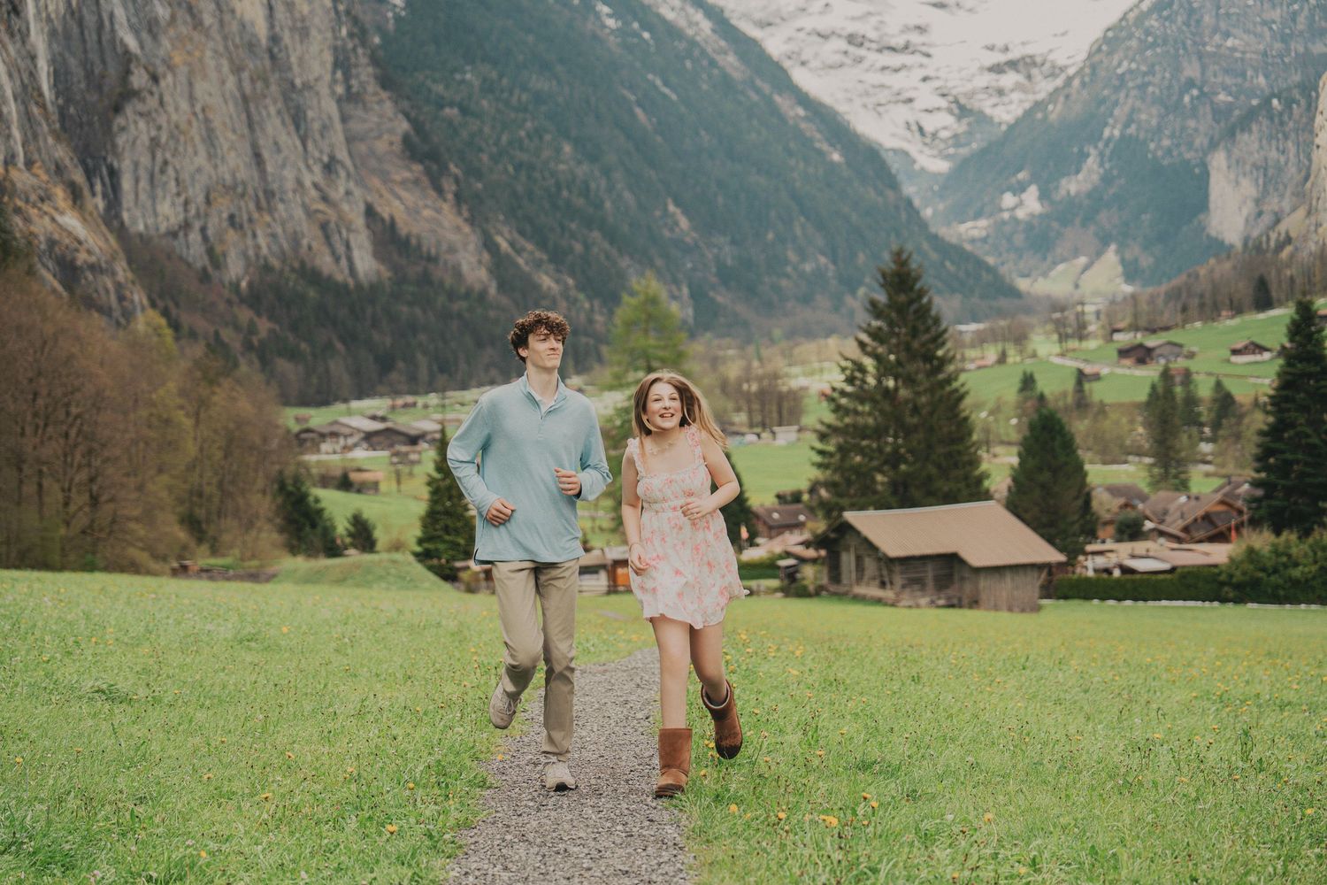 Joyful couple running through a green meadow with stunning Swiss Alps, evergreen trees, and rustic chalets behind them.