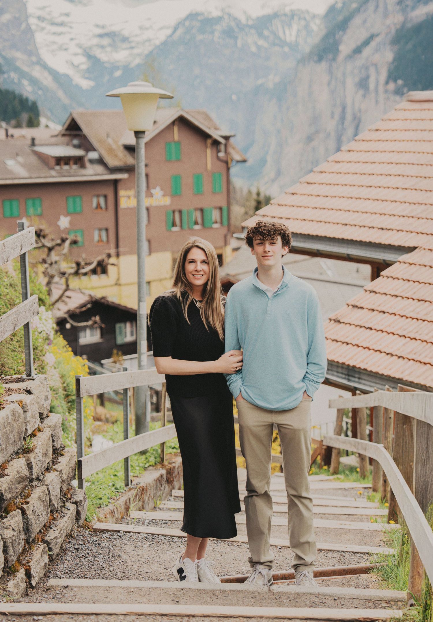 Mother in black dress and teenage son in blue sweater posing on stone steps in Swiss alpine village.
