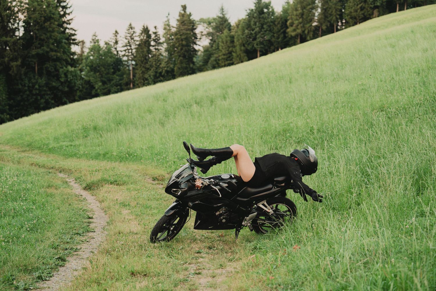 Person draped over black sport motorcycle on green grassy slope with forest backdrop.