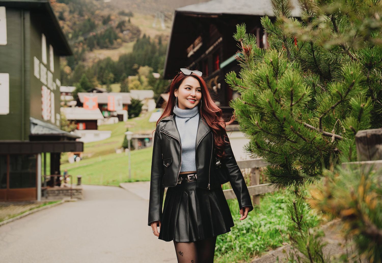 Smiling redhead woman in black leather jacket and skirt walking near pine trees in a mountain village.