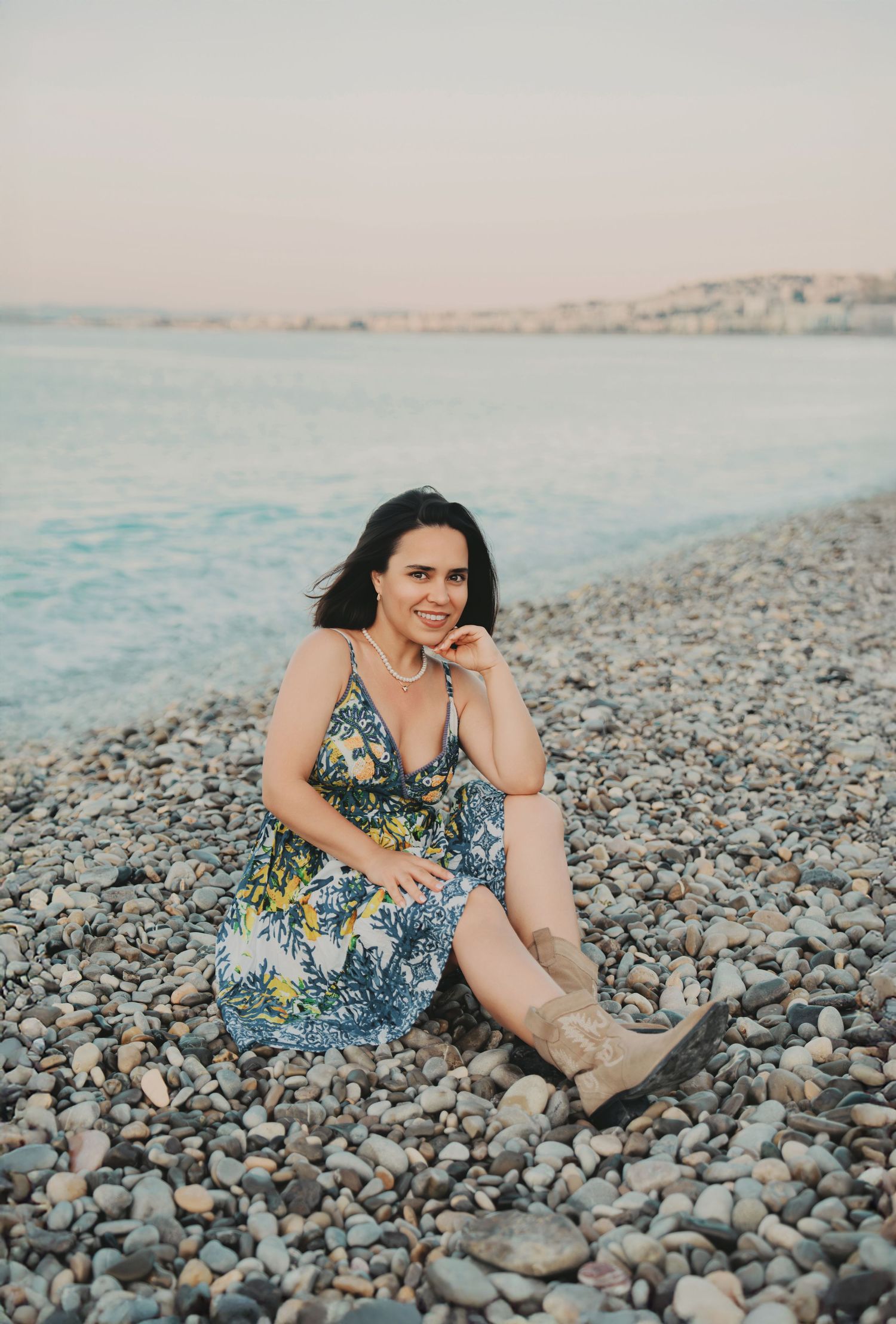 A woman in a floral sundress and boots sits smiling on a rocky seaside beach.