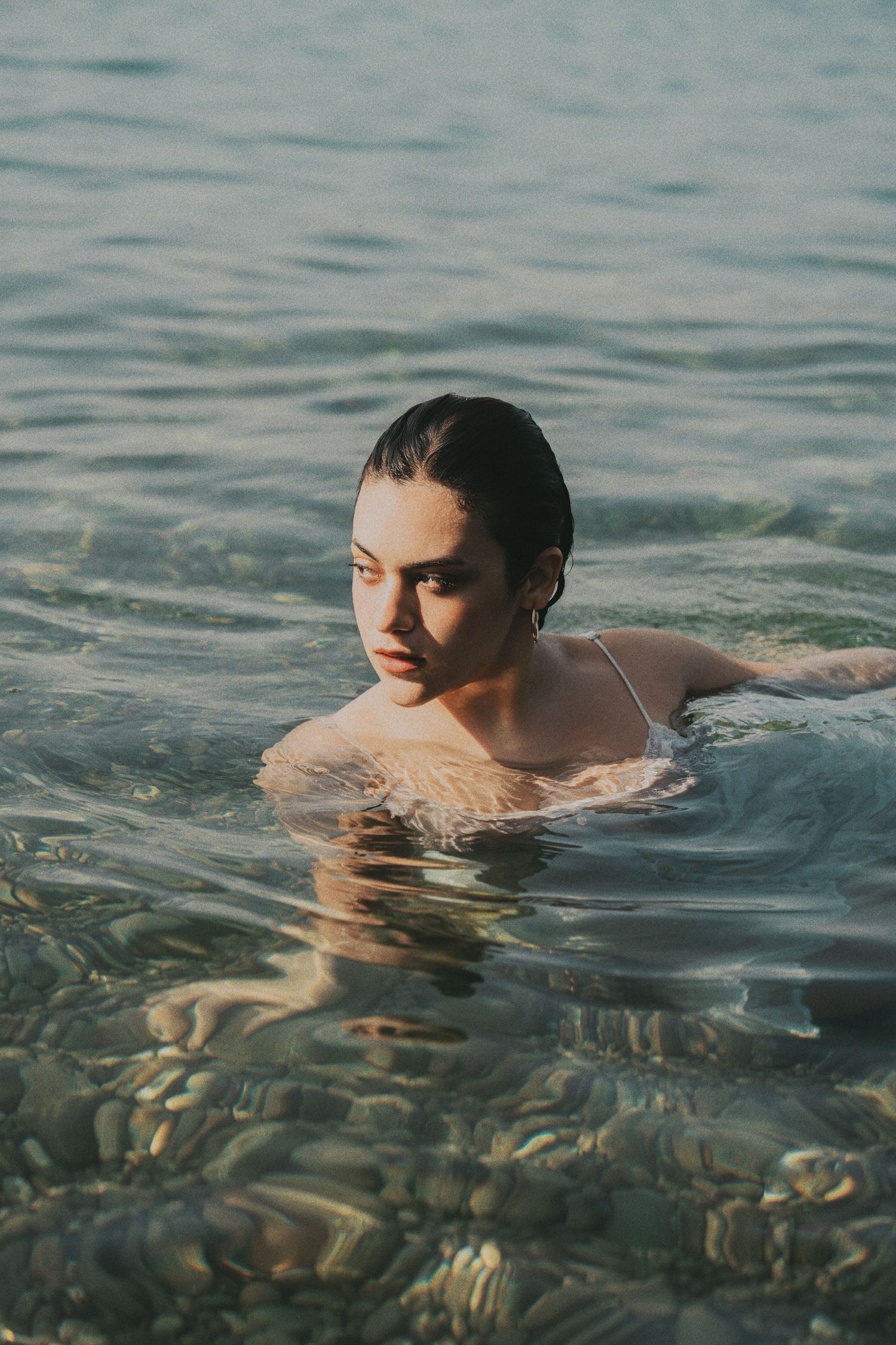 Young woman with slicked back wet hair swimming in clear shallow ocean water near rocky shore.