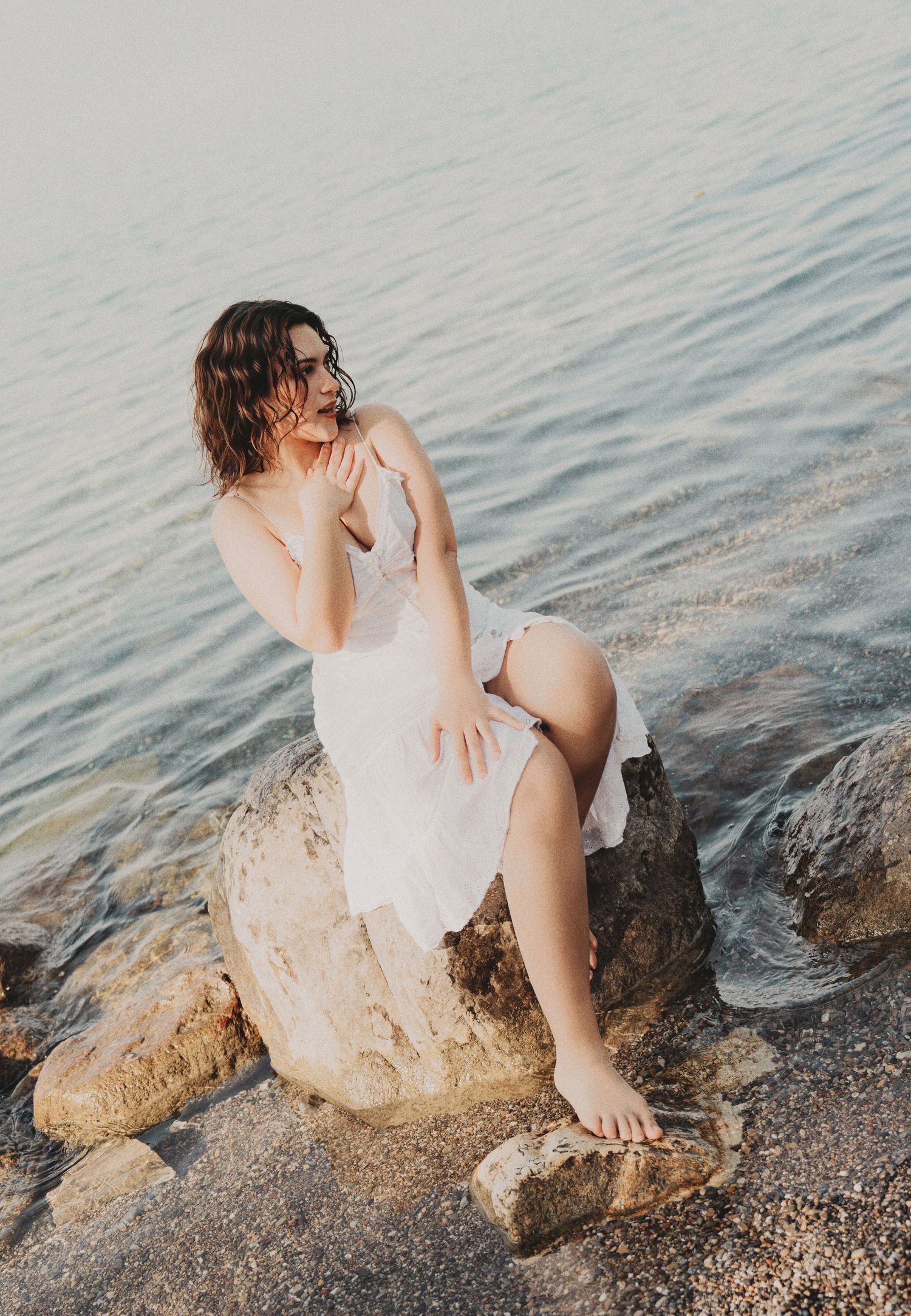 Woman in white dress sitting on coastal rocks by calm water at sunset.