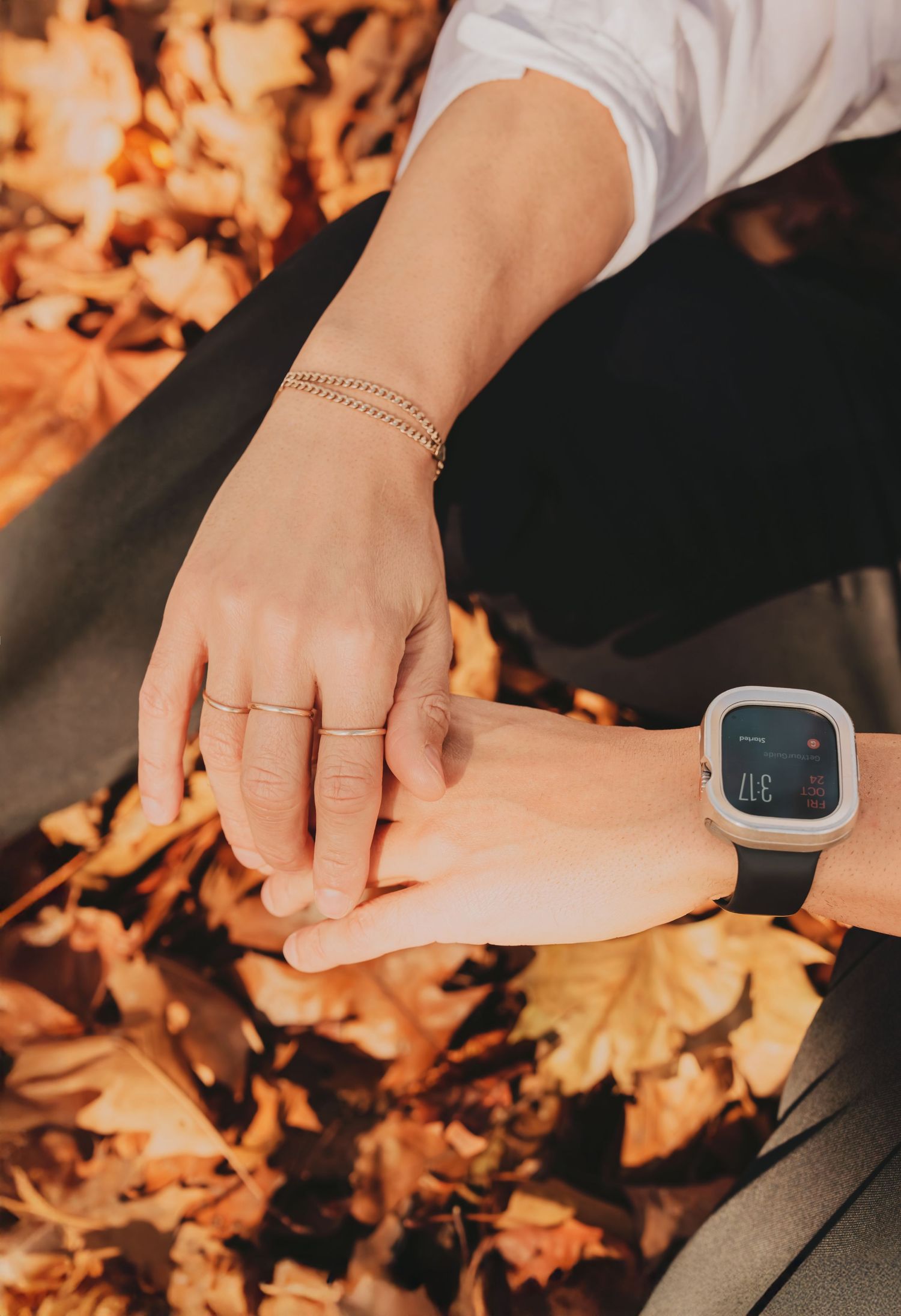 Close-up of two hands touching over autumn leaves, one wearing gold bracelet and the other a smartwatch.