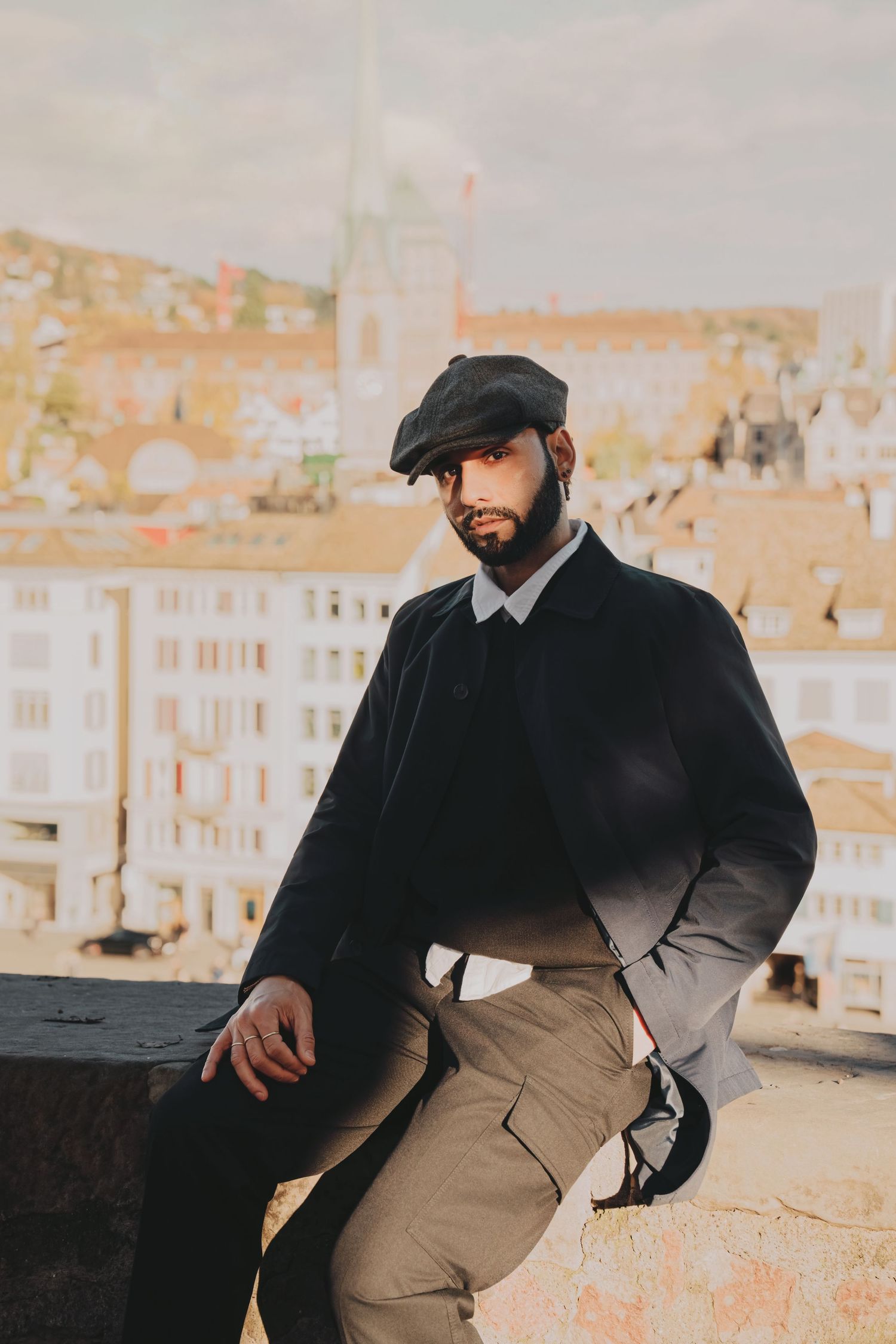 Bearded man in black jacket and flat cap sitting on a wall with a European cityscape in the background.
