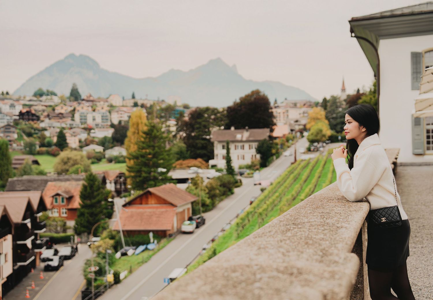 Woman in white sweater stands on balcony overlooking scenic European mountain town with misty peaks.