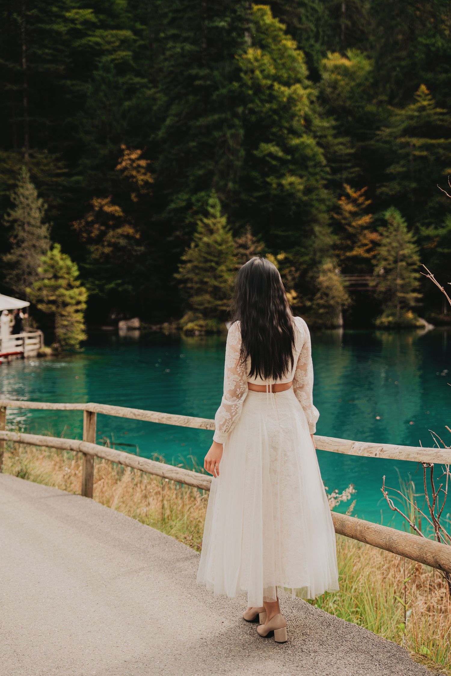 Woman in white lace top and tulle skirt standing with back turned overlooking a turquoise forest lake.