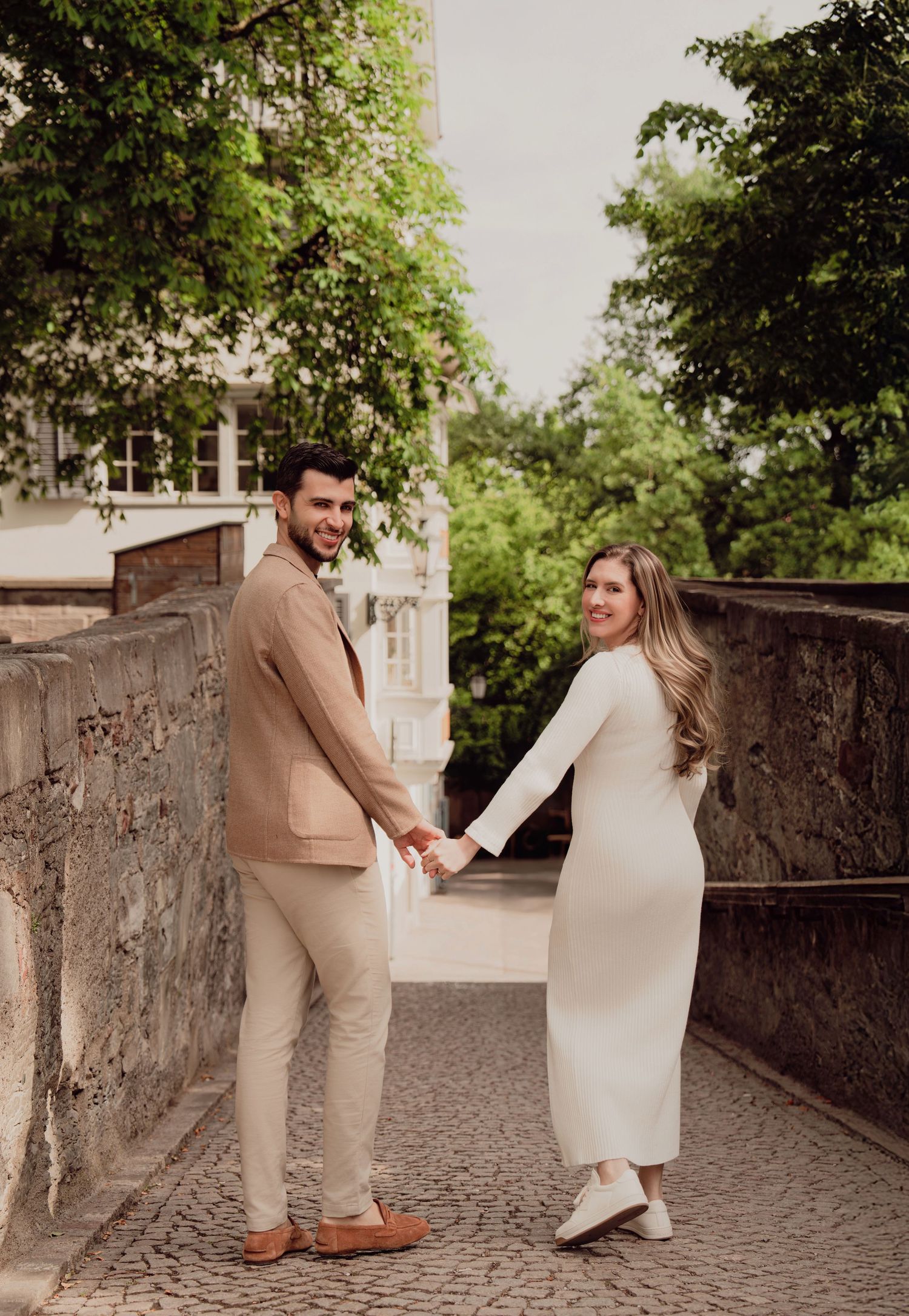 Expecting couple holds hands and looks back smiling while walking on cobblestone path in Zurich old town.