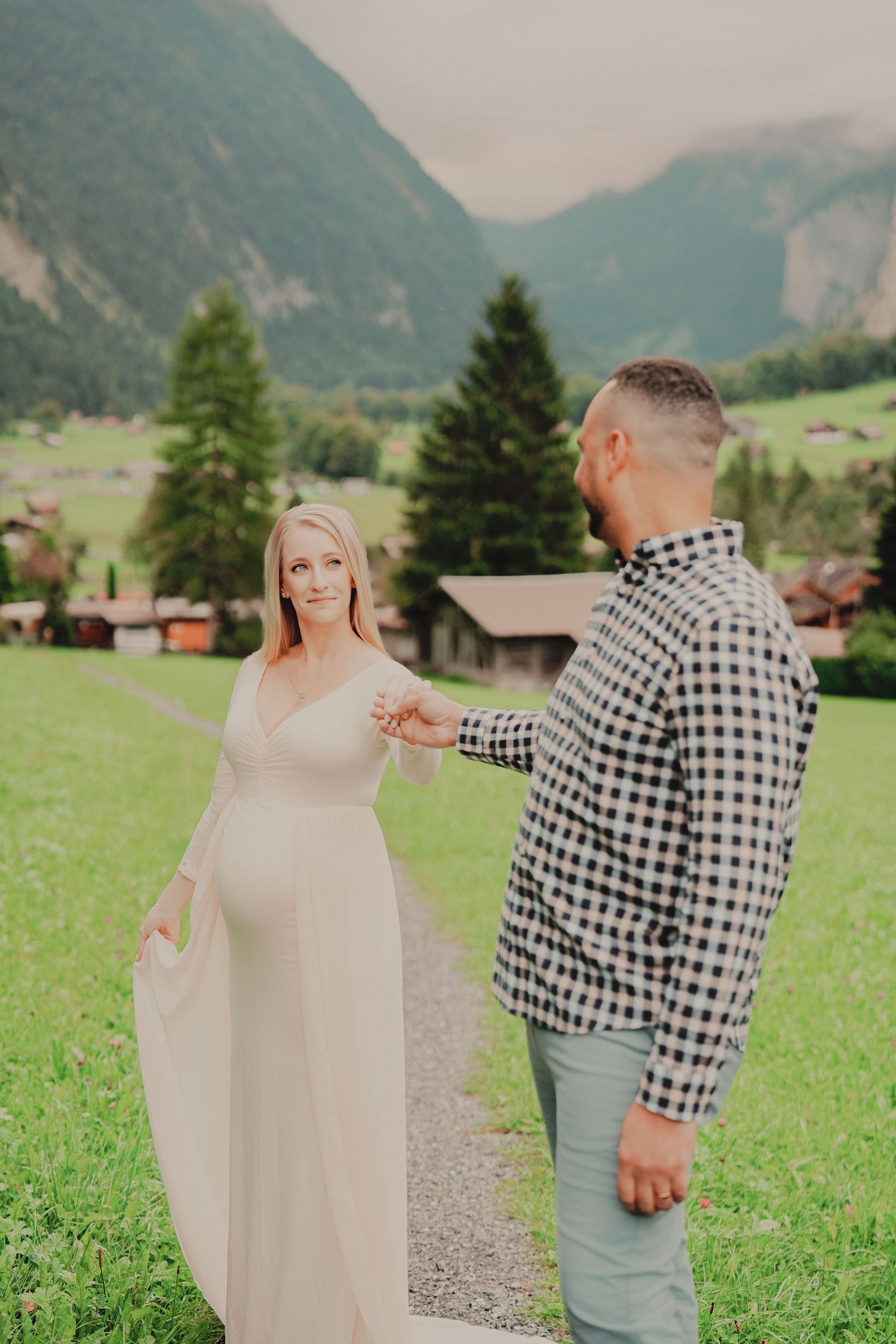 Pregnant woman in white dress holds hands with man in plaid shirt on mountain path in Swiss Alps.