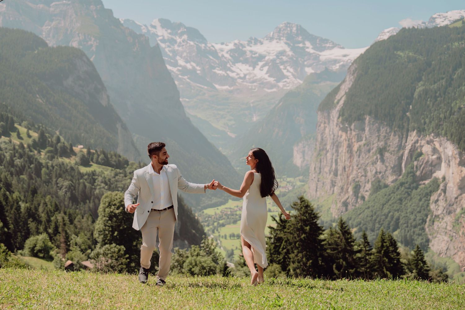 Couple holding hands walking through a green Alpine meadow with dramatic Swiss mountain valley views.
