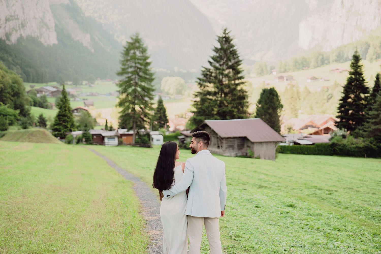 Bride and groom walk hand in hand through a green Alpine meadow with mountain village in the background.