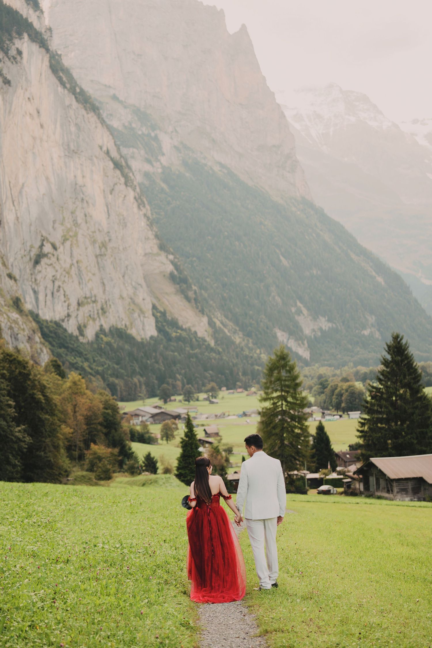 Couple walking hand-in-hand on mountain path with dramatic Swiss Alps cliffs behind them.