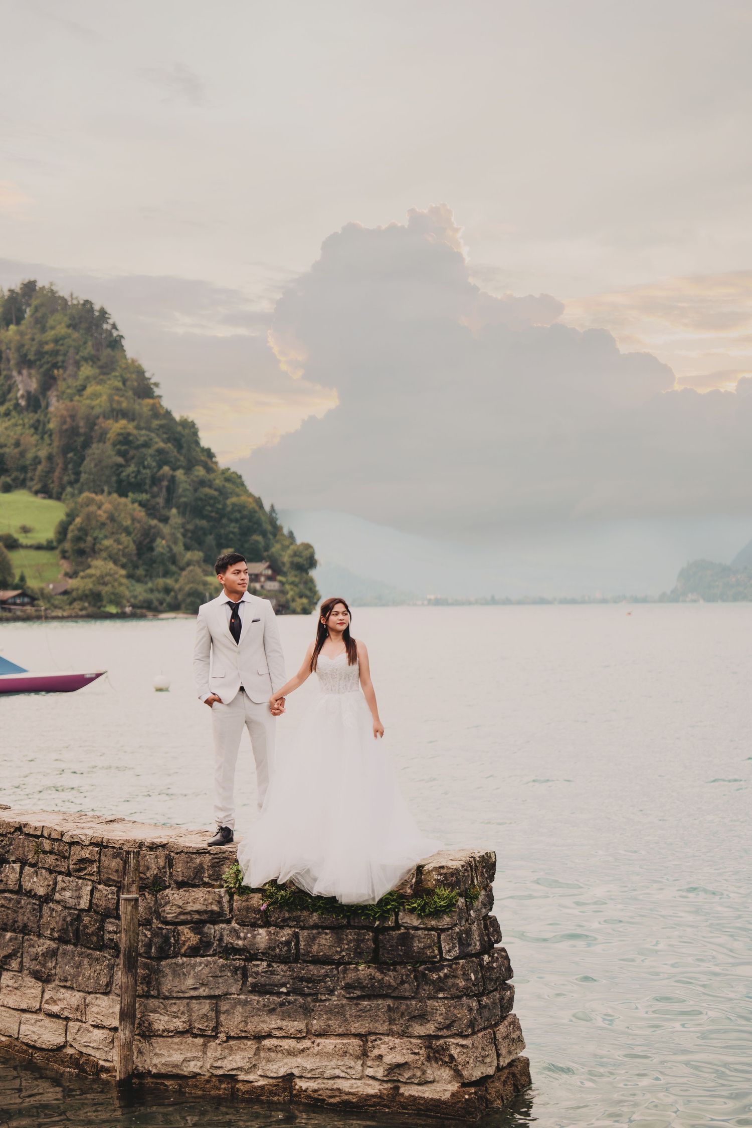 Wedding couple holding hands on stone jetty with misty alpine lake and forested mountain behind.