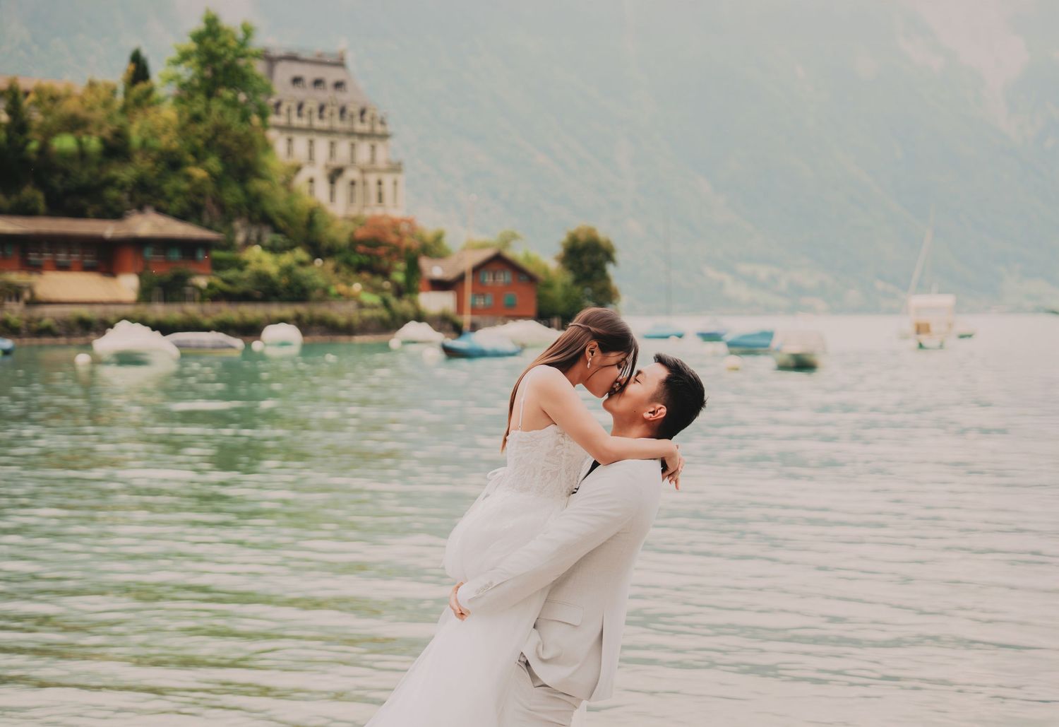 Groom lifting bride in a romantic embrace by a scenic alpine lake with boats and mountains behind them.