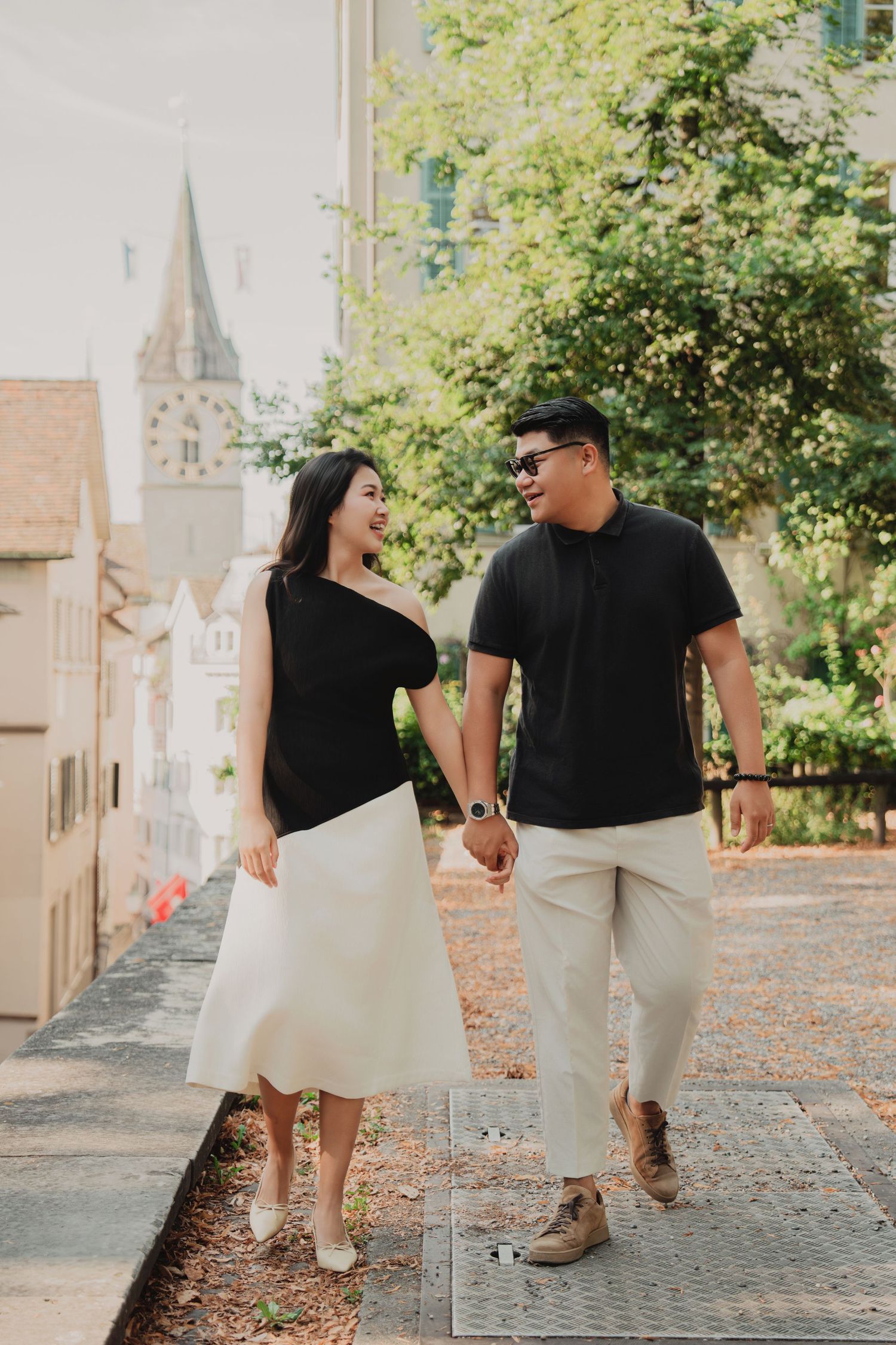 A couple holds hands and walks together through a charming European street, smiling at each other.