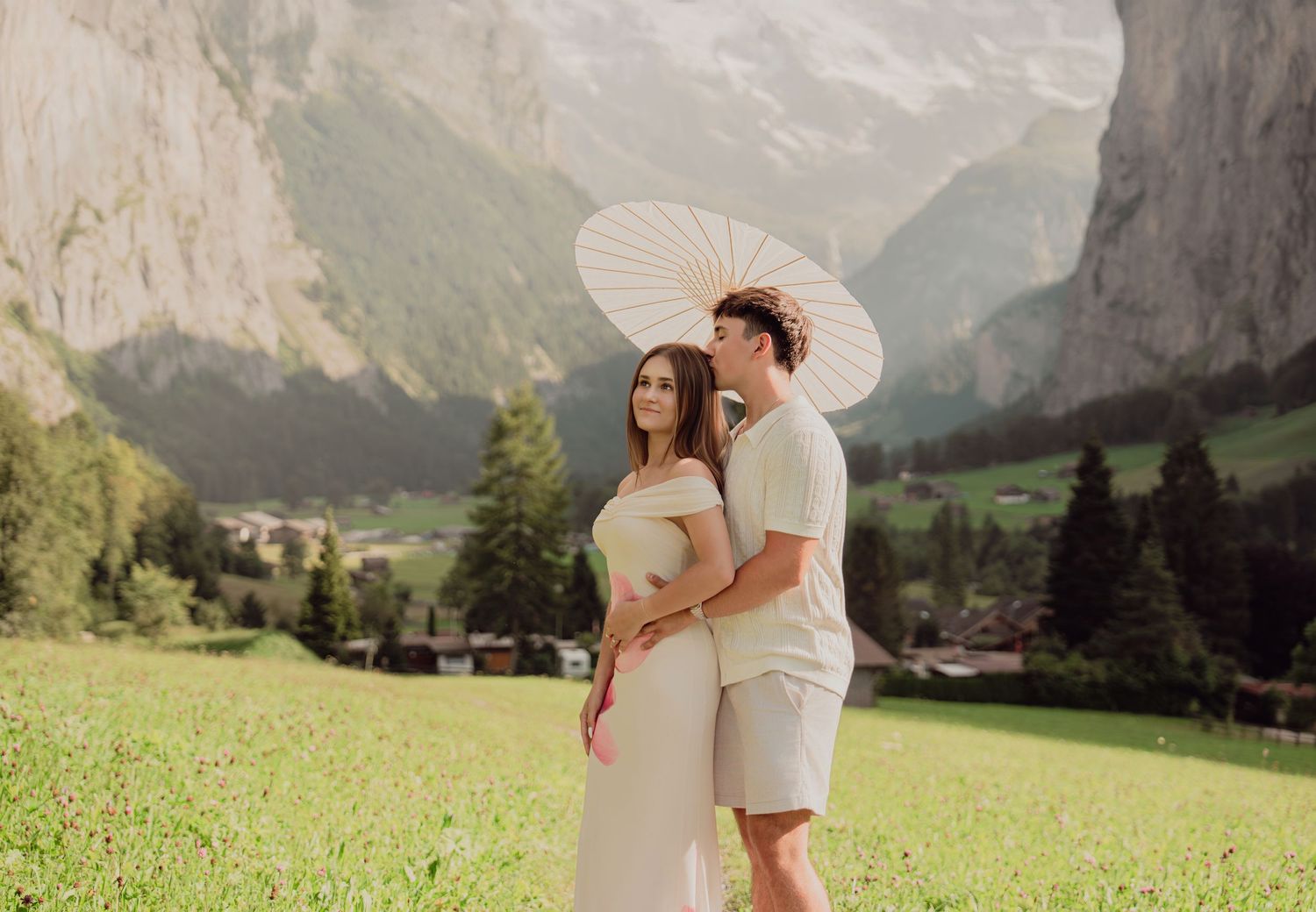 Elegant couple posing with a white parasol in a scenic Swiss alpine meadow with dramatic mountain cliffs behind them.