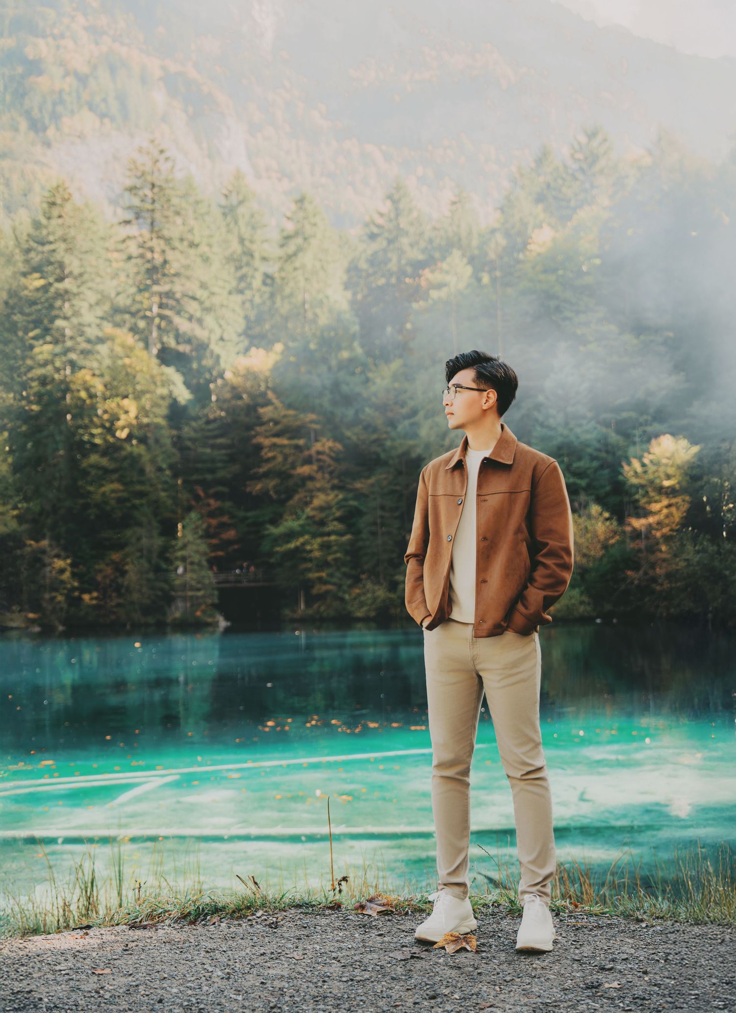 Stylish man in brown jacket standing before a vivid turquoise lake with forested mountains.