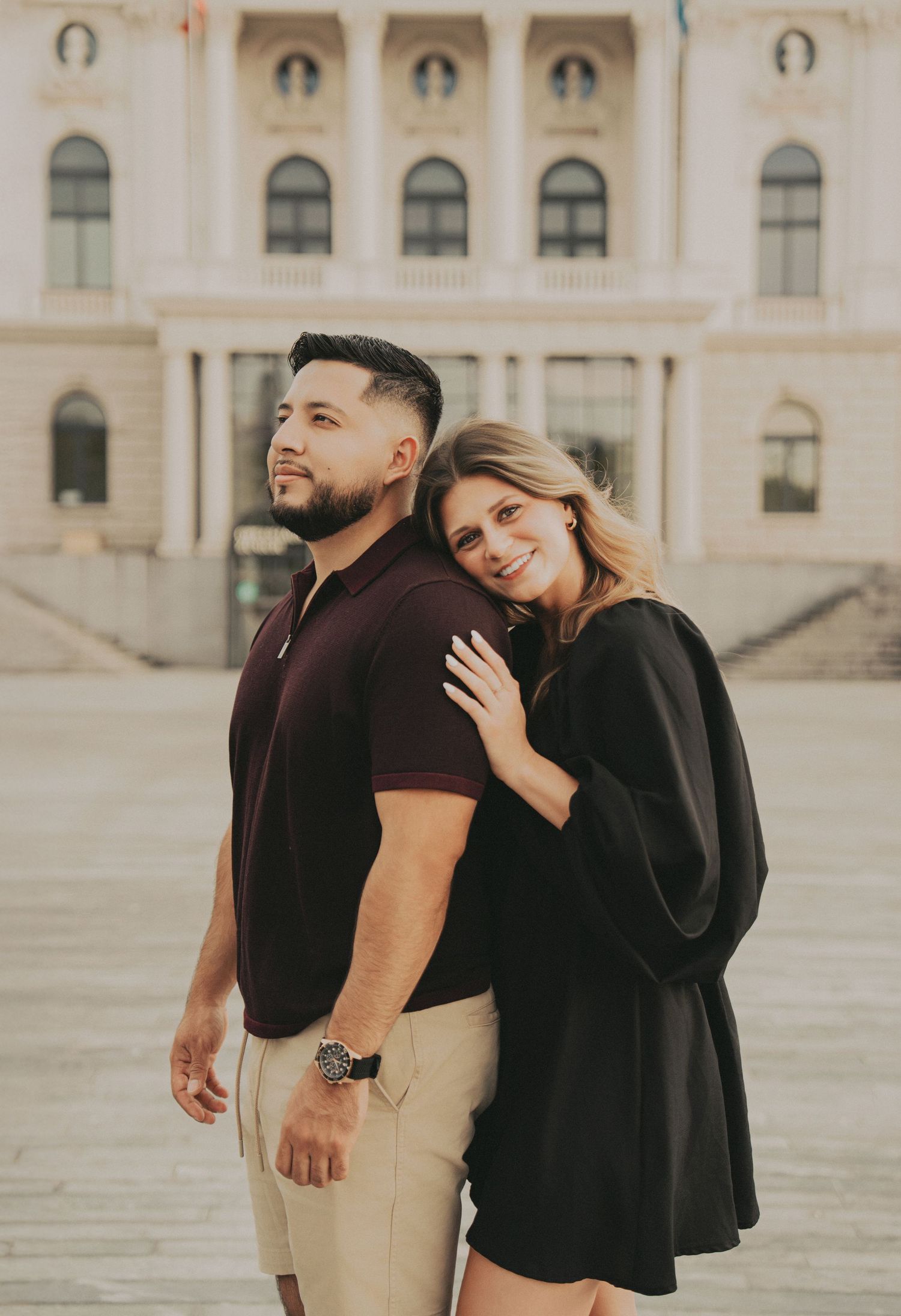 Couple posing back to back with woman smiling and leaning on mans shoulder in a European plaza.