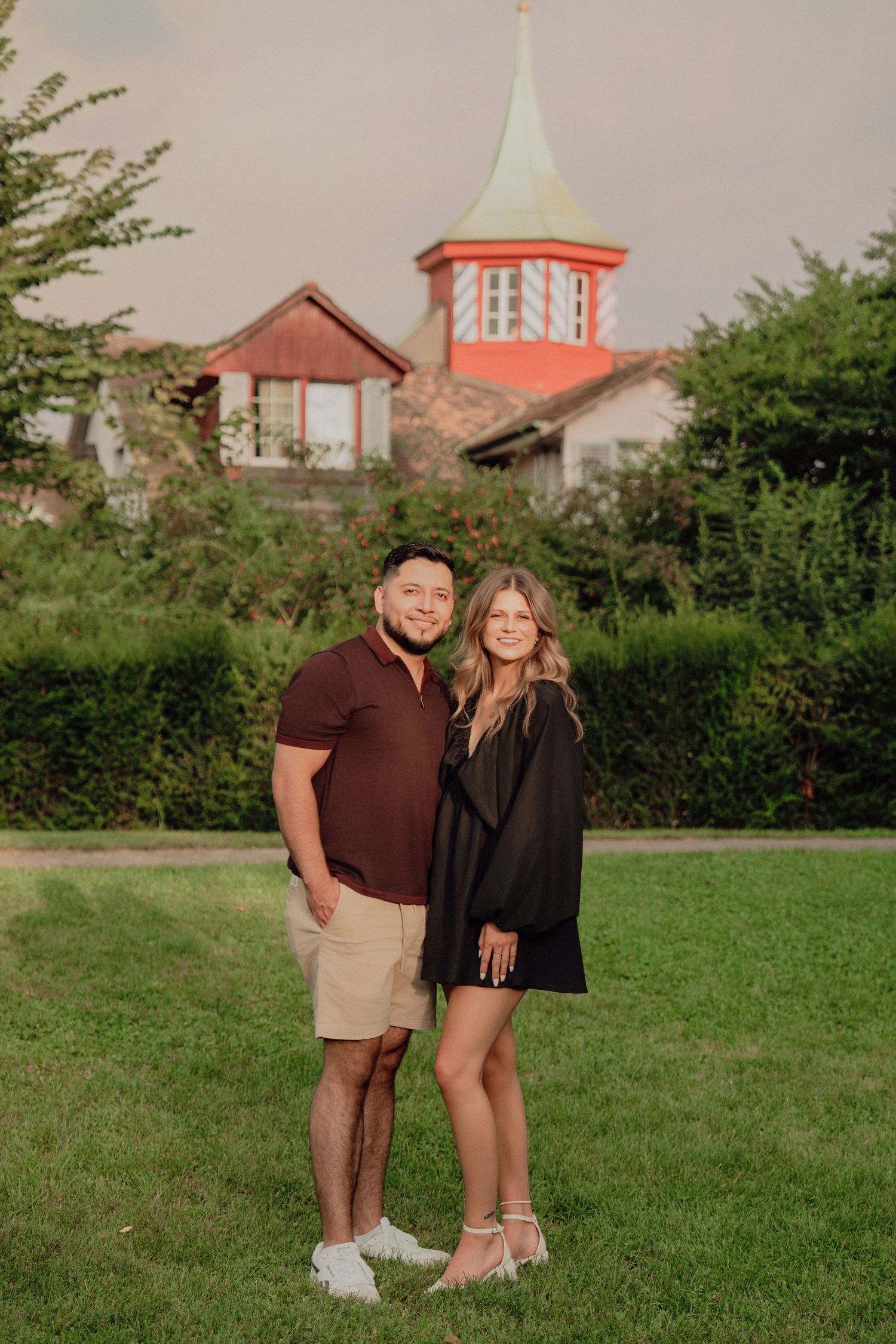 Happy couple standing together on a manicured lawn with a distinctive red-topped turret building behind them.