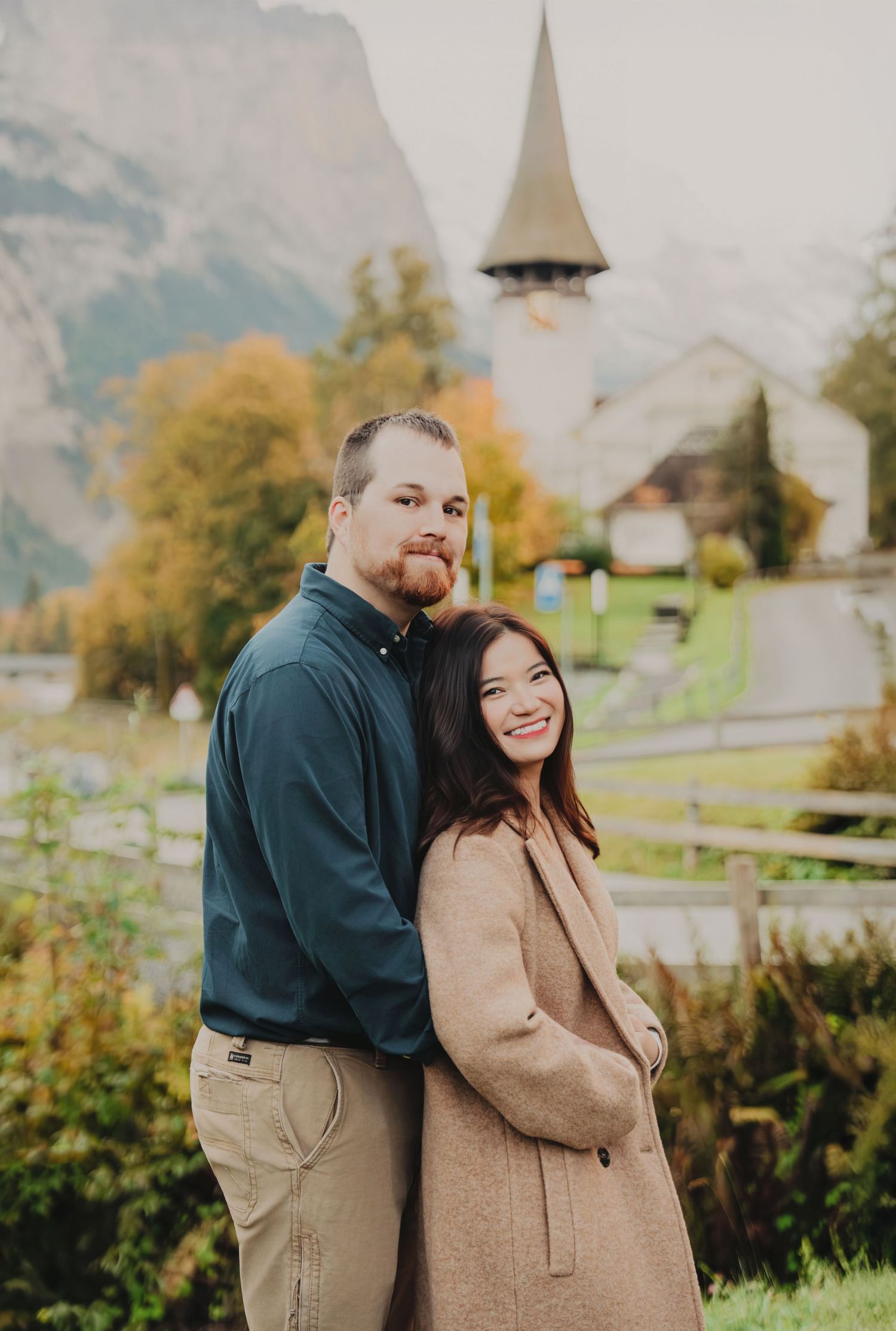Couple posing back-to-back near a historic Swiss church steeple surrounded by autumn foliage and mountains.