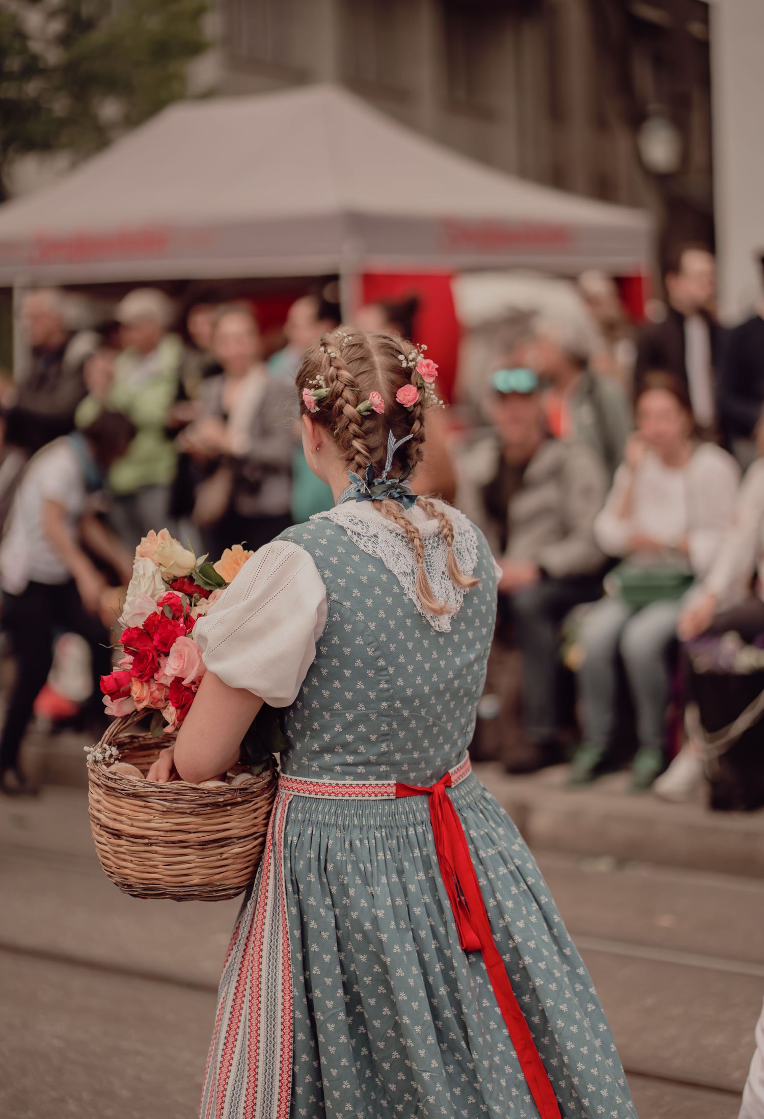 Girl in traditional dirndl dress with braided hair carries flower basket at cultural festival parade.