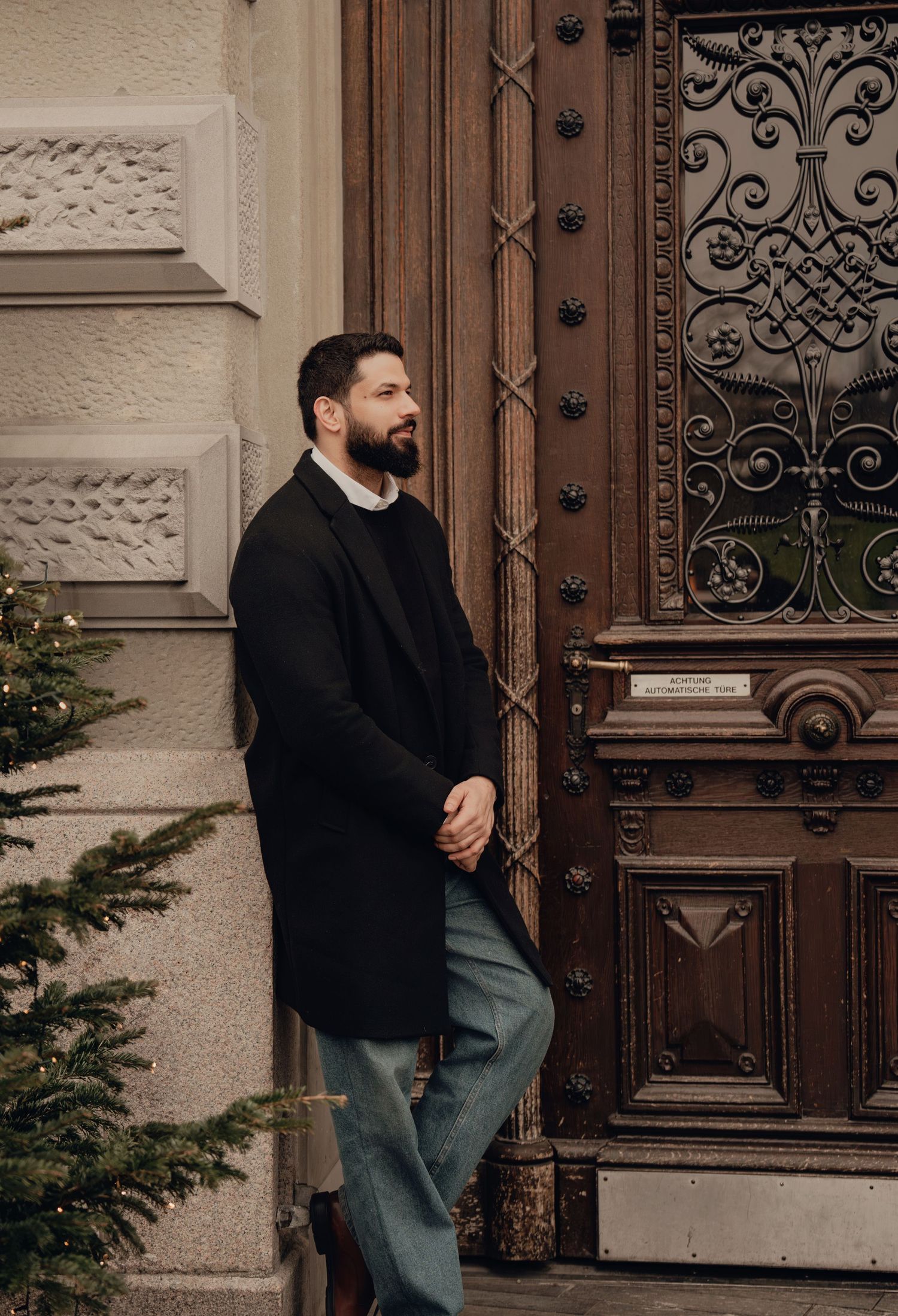Bearded man in black coat leaning against ornate wooden door with wrought iron details beside Christmas tree.