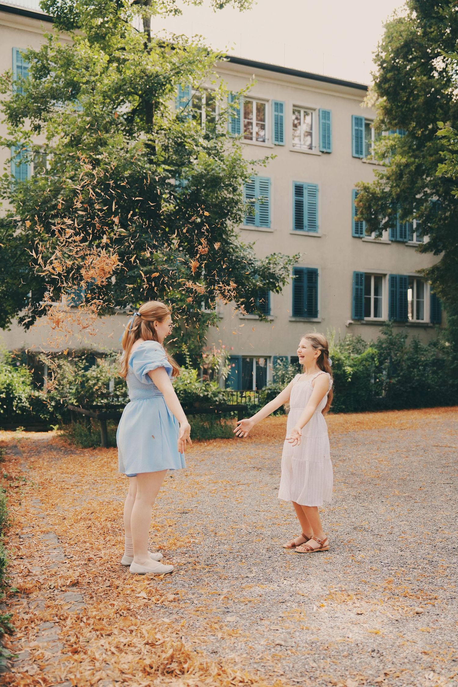 Two girls in blue and white dresses hold hands in a sunlit courtyard near a European building.