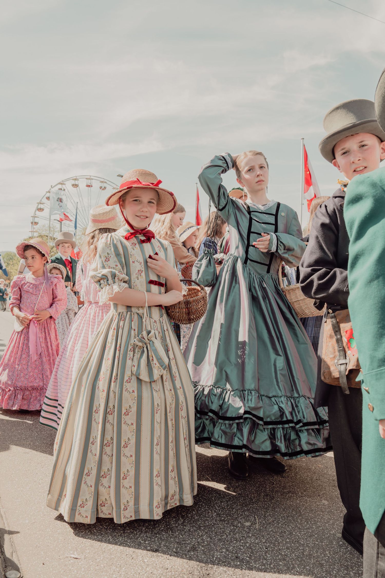 Children in Victorian era costumes parade at outdoor festival with ferris wheel backdrop.