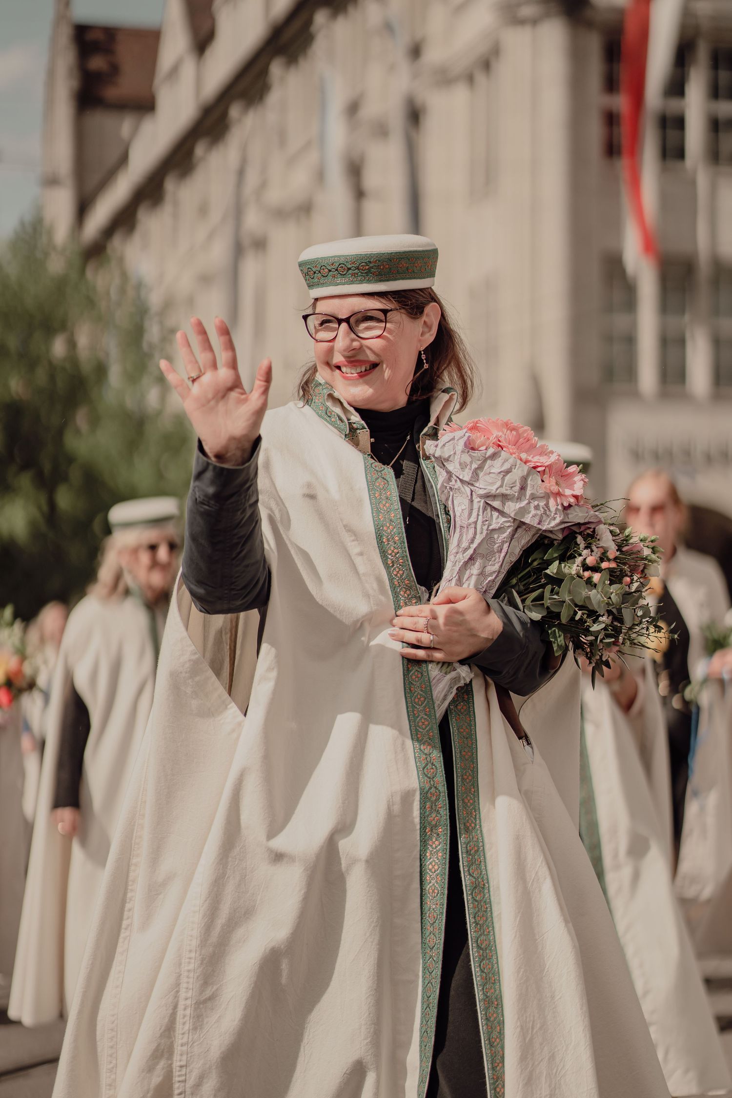 Smiling woman in white ceremonial robe waves while holding pink flower bouquet at parade.