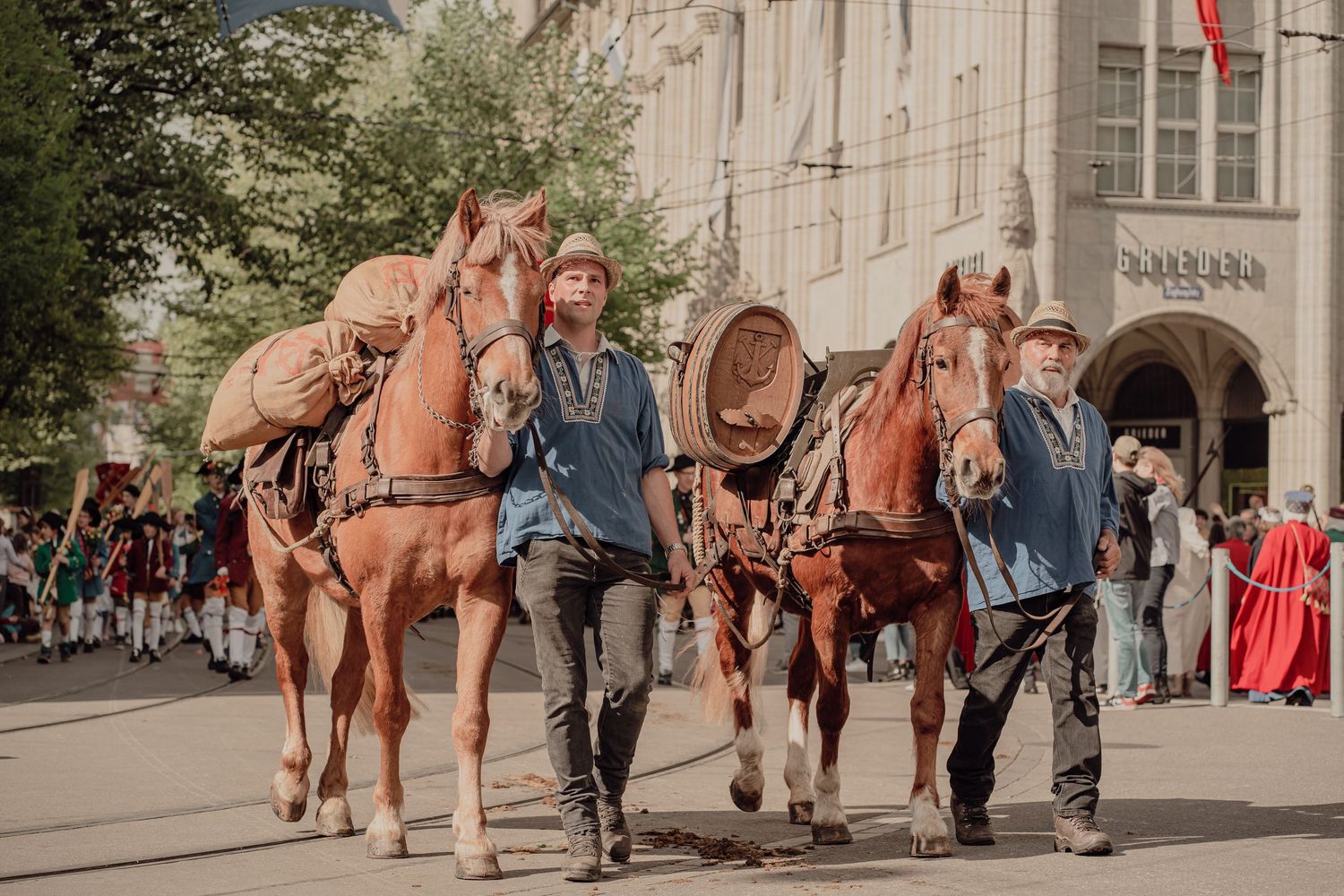 Two men in medieval blue tunics lead chestnut horses through a crowded historical street parade.