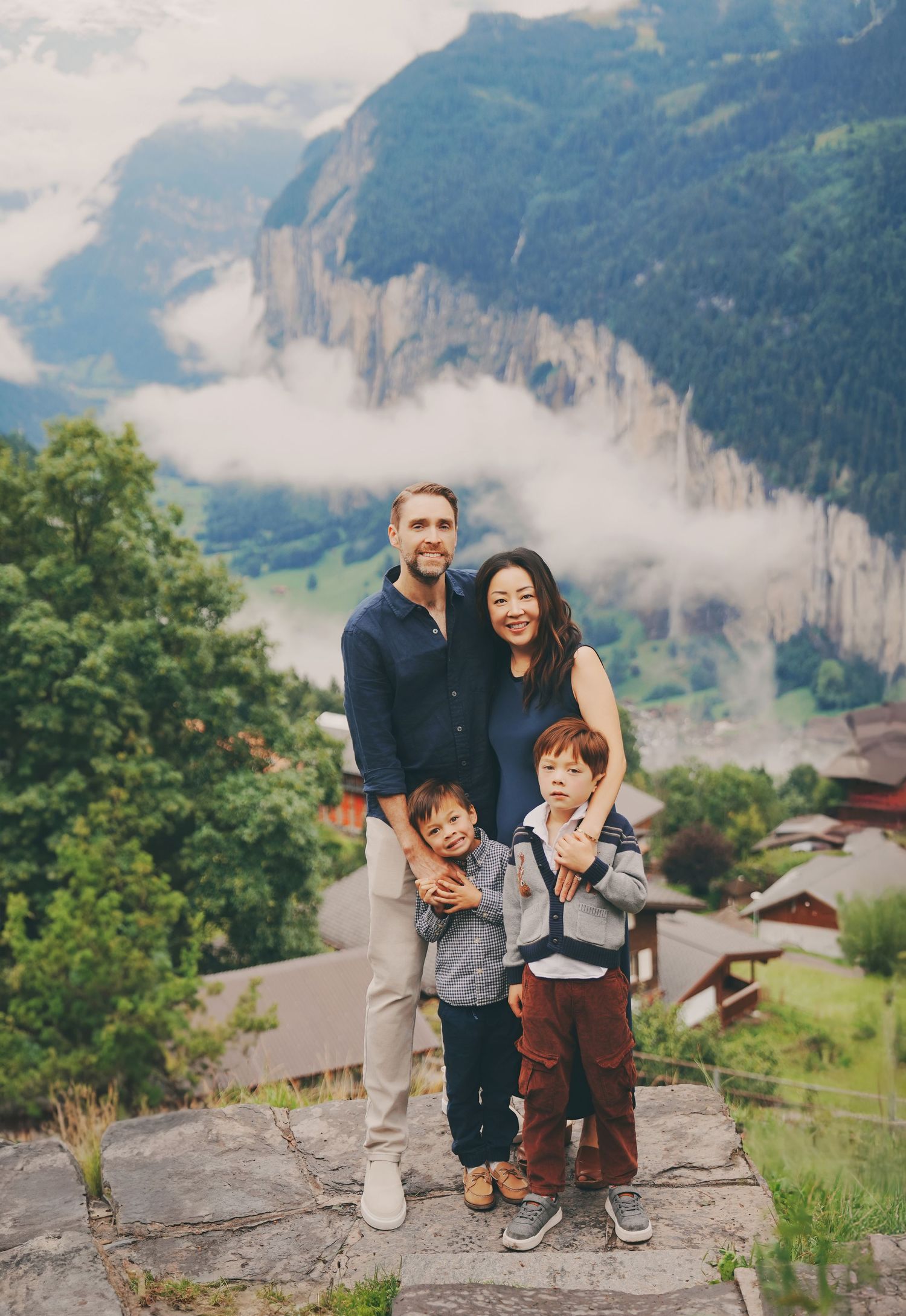 Smiling family with two young boys stands on stone ledge with misty Swiss Alpine valley backdrop.