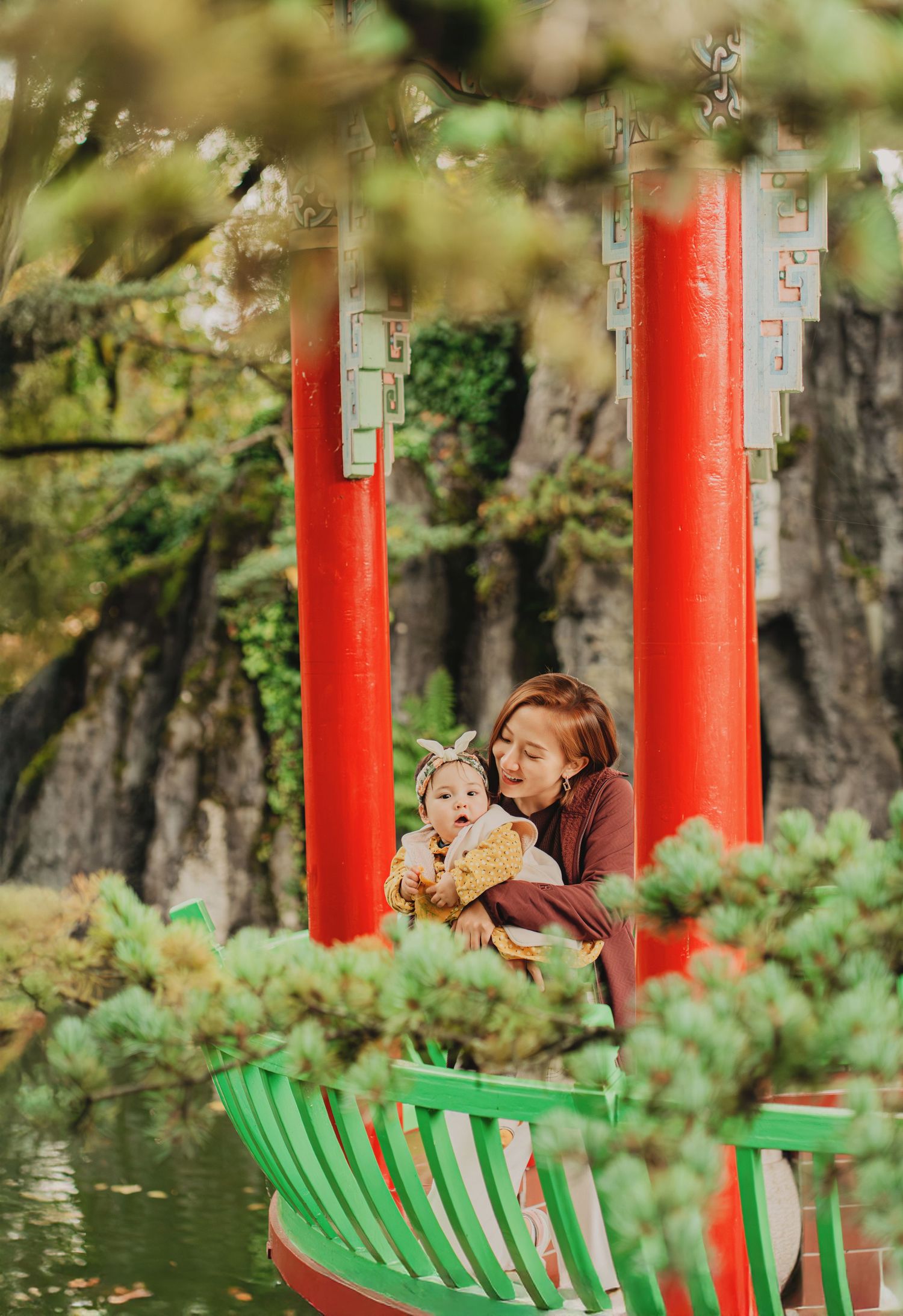 Young mother tenderly holding laughing baby near vibrant red pillars in Chinese garden.