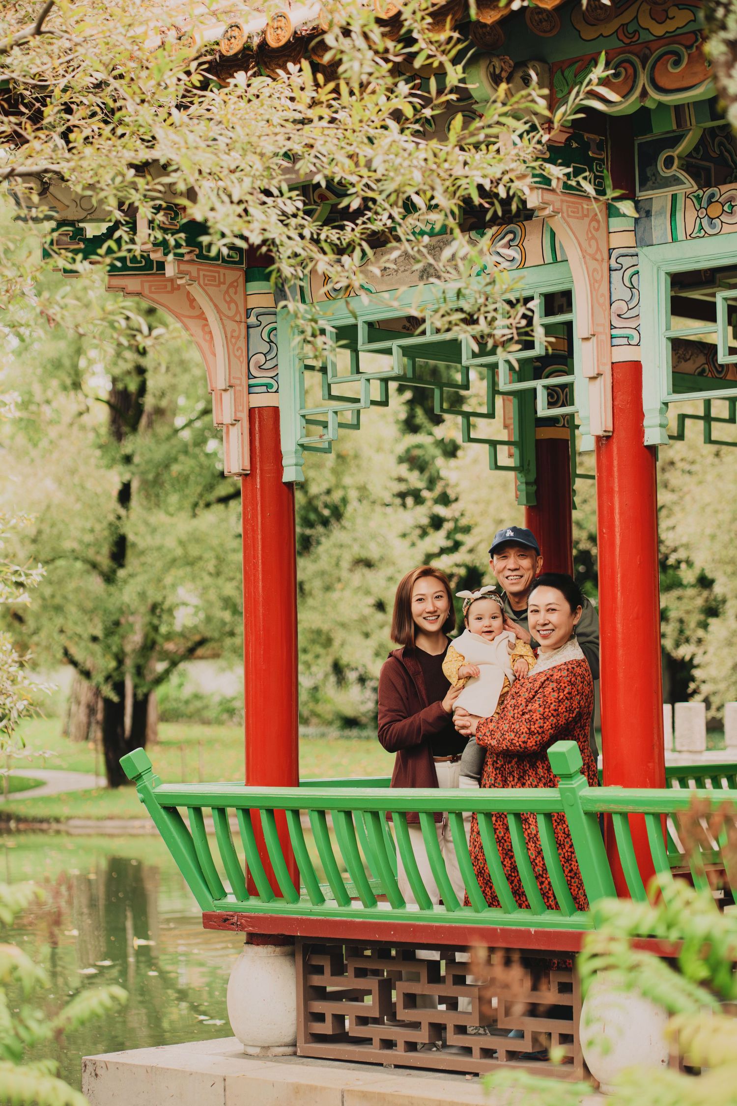 Happy family with baby posing inside colorful traditional Chinese pavilion with red columns beside a pond.