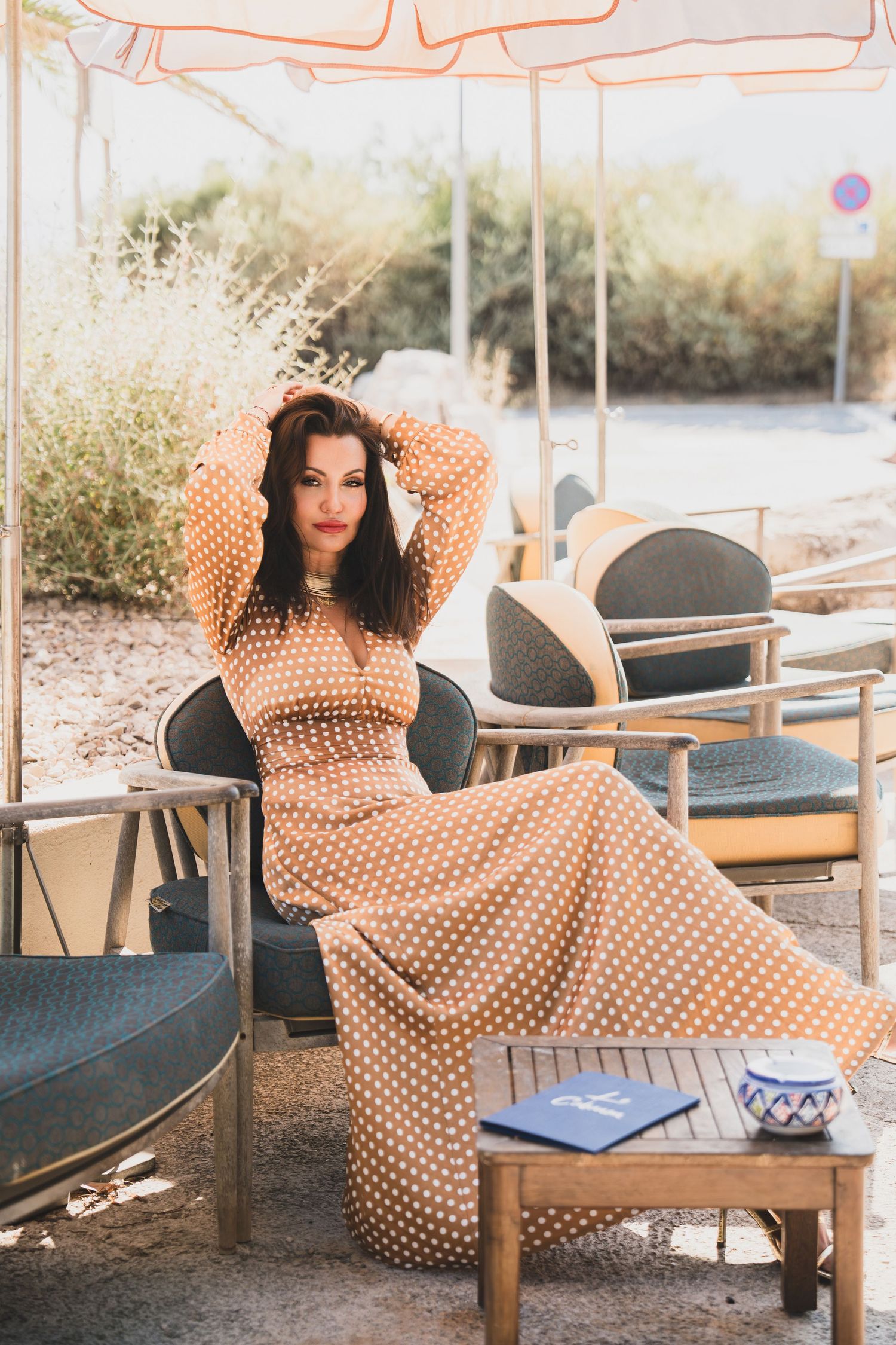 Woman in polka dot dress posing in outdoor cafe seating area surrounded by natural greenery.