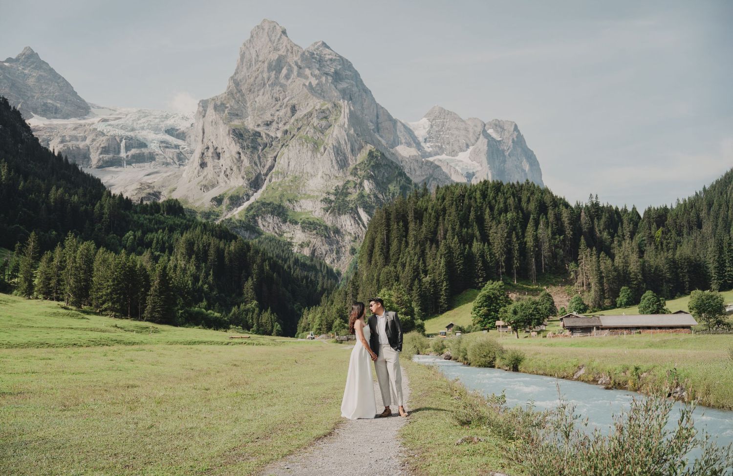 Bride and groom holding hands on a scenic path with snow-capped peaks and green valley.