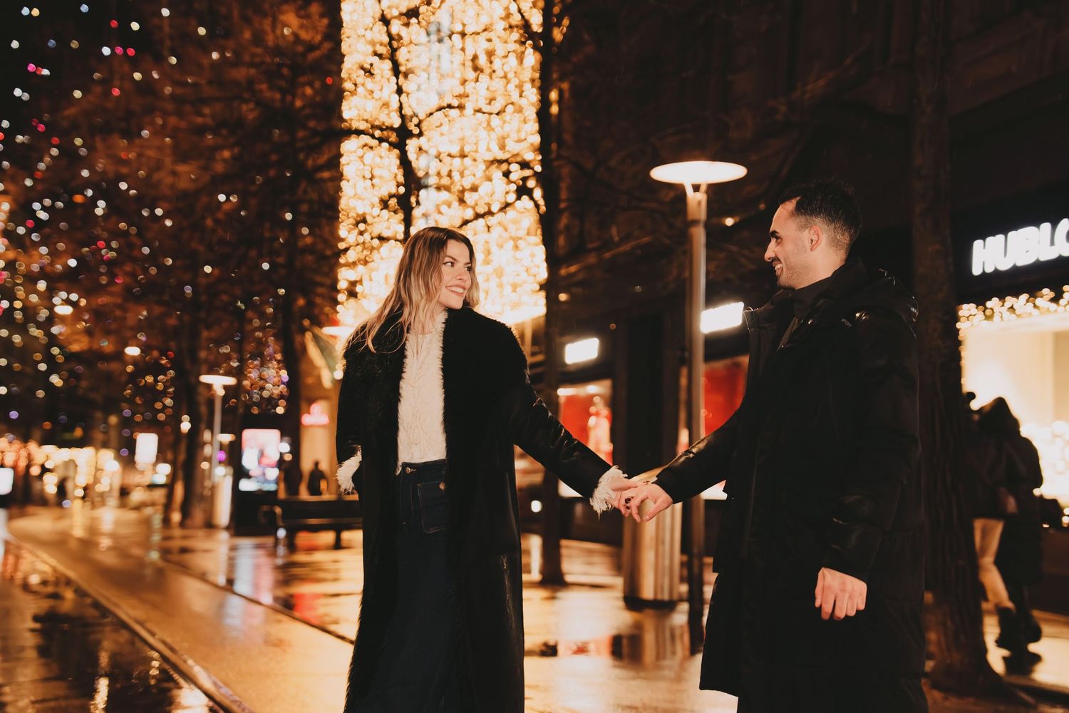 Couple holding hands on a rain-slicked city street adorned with beautiful Christmas lights at night.