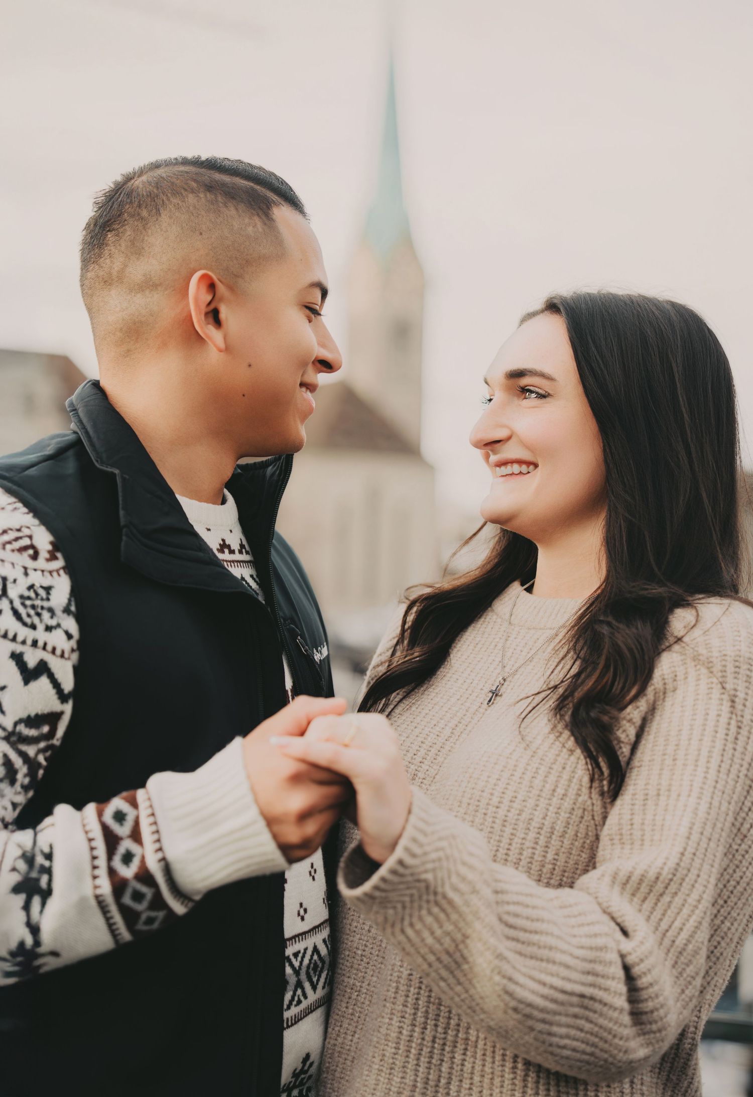Smiling couple gazing at each other holding hands with church steeple in Zurich backdrop.