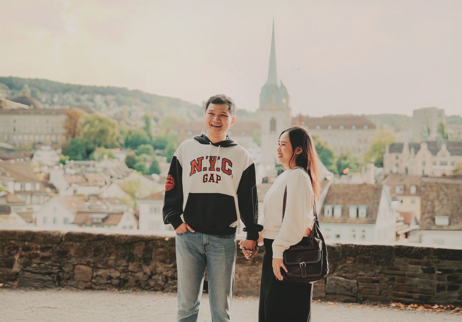 Happy couple holding hands on a scenic overlook with Bern old town rooftops and church spire behind them.