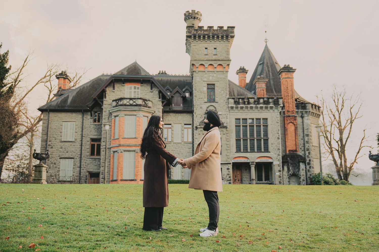 Couple holding hands before grand historic castle on cloudy day.