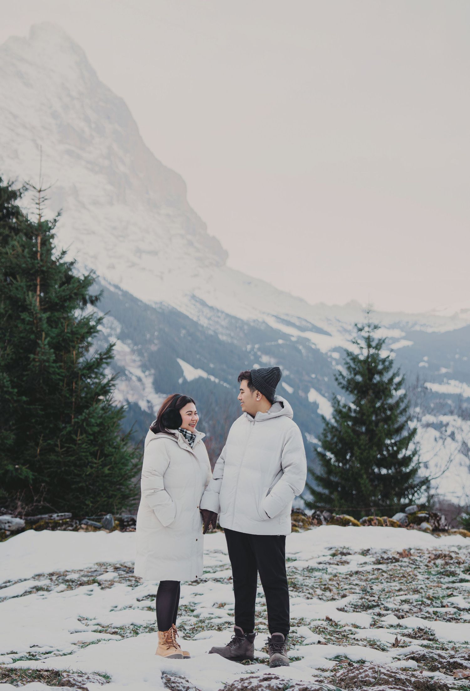 Couple in matching white puffer coats standing in snowy alpine landscape with mountains and evergreen trees.